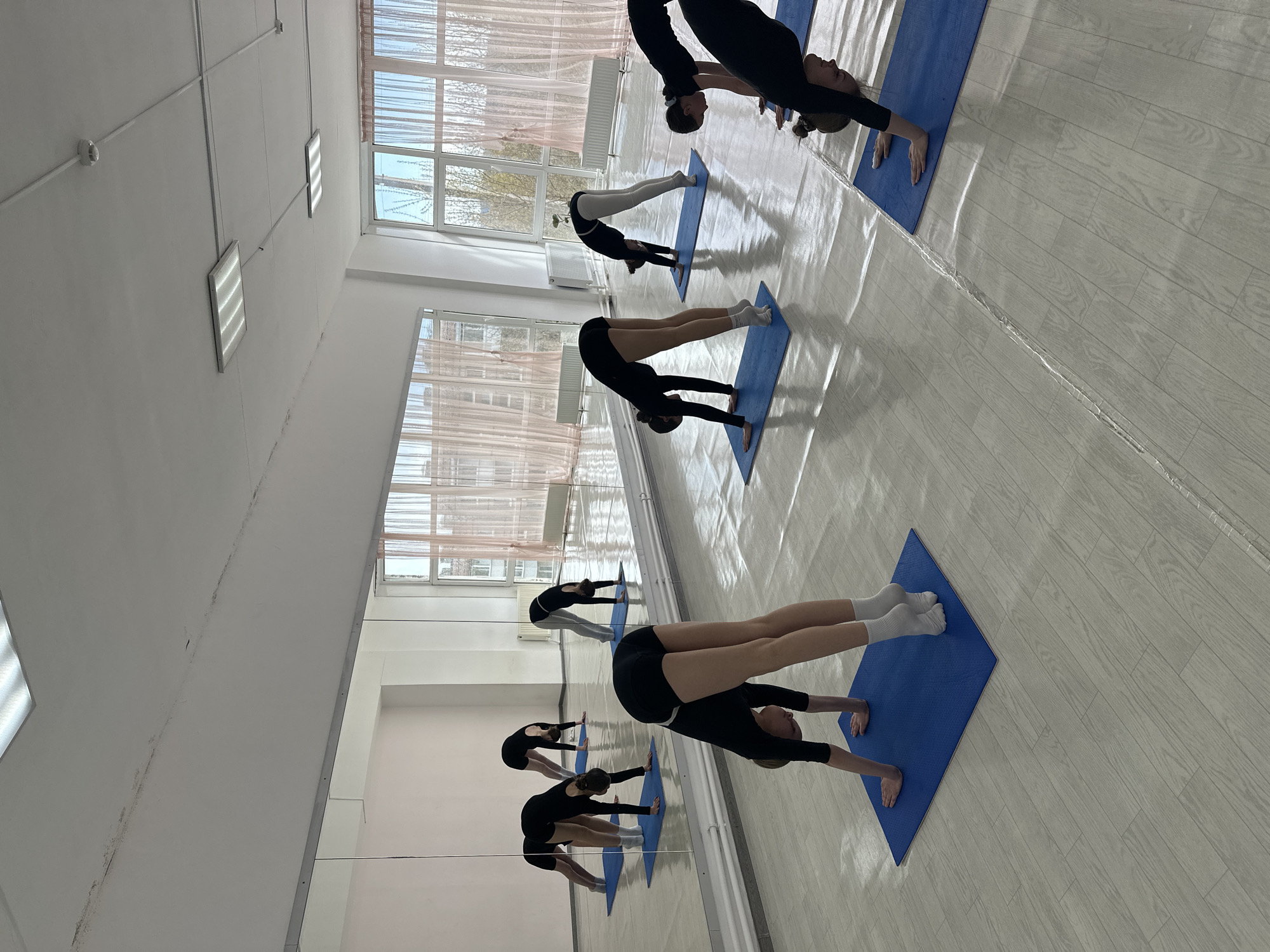 This image depicts an indoor exercise or dance studio in Votkinsk, Russia, during what appears to be daytime. Several young women, dressed in black athletic wear, are engaged in a stretching or yoga session. They are performing an inverted V-shaped pose, similar to a downward dog, with their hands and feet placed on blue exercise mats.
The studio features a long mirrored wall on the left side, which reflects the participants and the room, creating an expanded visual of the space and the activity. Opposite the mirror, large windows with light-colored, sheer curtains allow natural light to illuminate the room. Through the windows, an outdoor view of trees, sky, and distant buildings is visible. White radiators are installed below the windows.
The ceiling is white and equipped with several rectangular fluorescent light panels, connected by visible conduit. The floor is covered with light-colored wooden laminate flooring, reflecting some of the overhead lighting. The overall scene conveys a sense of focused physical activity within a bright and functional studio environment. No discernible text is visible in the image.