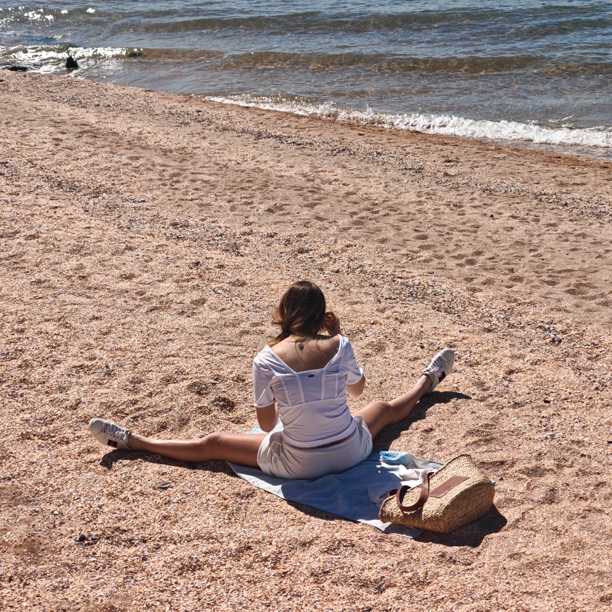 A woman sits on a beach with her legs in a full split, wearing a white top, white shorts, and white sneakers. She is on a blue towel, with a straw bag beside her. The beach is made of sand and small shells, and the ocean is in the background. The location is unknown.