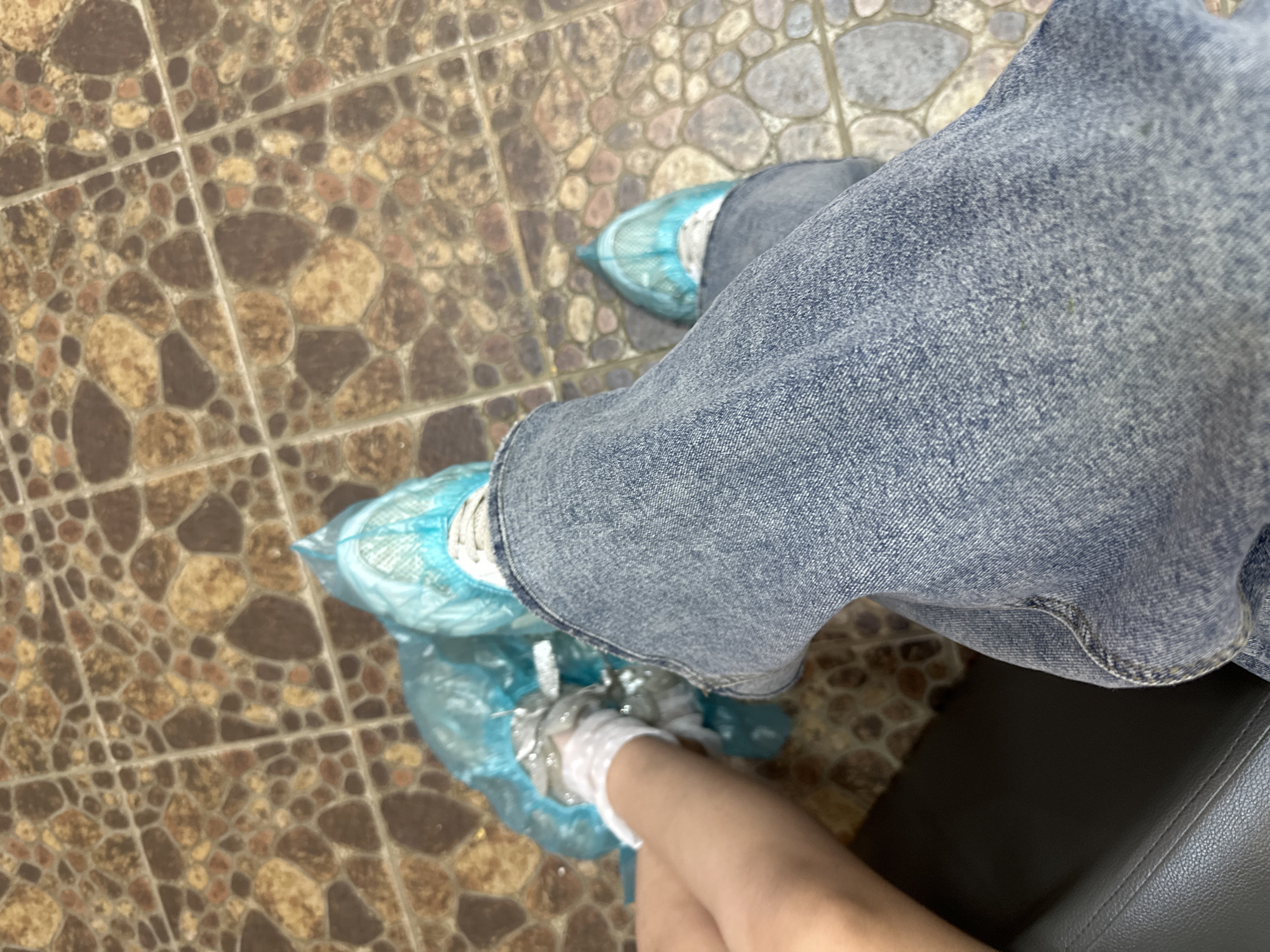 A view from above shows a person seated, wearing blue jeans and disposable blue plastic shoe covers over their feet, on a floor with a pattern of small, irregularly shaped brown and tan tiles. Part of a dark grey chair or bench is visible on the right. The location is Unknown, Unknown.