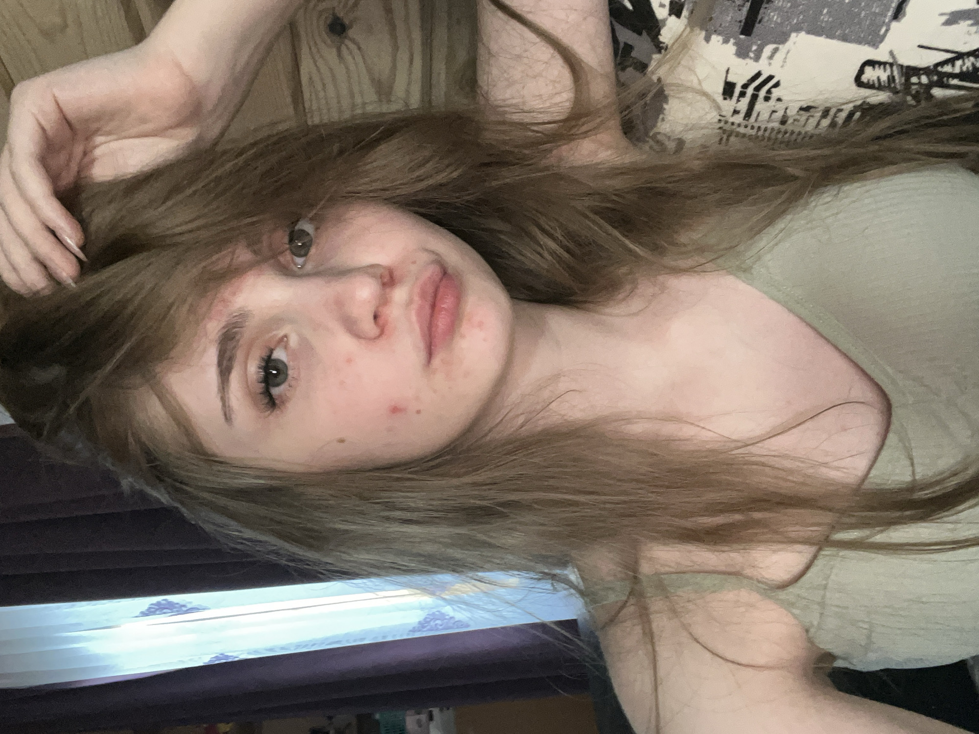 The media file is a close-up, rotated selfie of a young person. The subject has long, light brown hair that is spread out around their face and shoulders. They have fair skin with some visible blemishes, including red spots on their cheeks and forehead, and freckles. Their eyes are light green, and they are looking towards the camera with a neutral or slightly content expression. Their lips are slightly parted. They are wearing a light green, ribbed top. One arm is raised and bent, with their hand resting on their head and hair. The background is a mix of textures: to the left, there appears to be a wooden surface with visible grain and knots. To the right, a patterned fabric, possibly a pillow or blanket, is visible, with abstract black and white designs. In the lower part of the image, there is a view of what looks like blinds or a window covering with horizontal stripes of light and darker colors, suggesting an indoor setting, possibly a bedroom. The lighting appears to be soft and coming from a source above and to the left, casting gentle shadows. There is no visible text in the image. Given the context provided, the location is Ashkadar, Russia. The time of day and weather are not discernible from the image alone. The activity is a selfie, likely taken for personal enjoyment or to share with others.