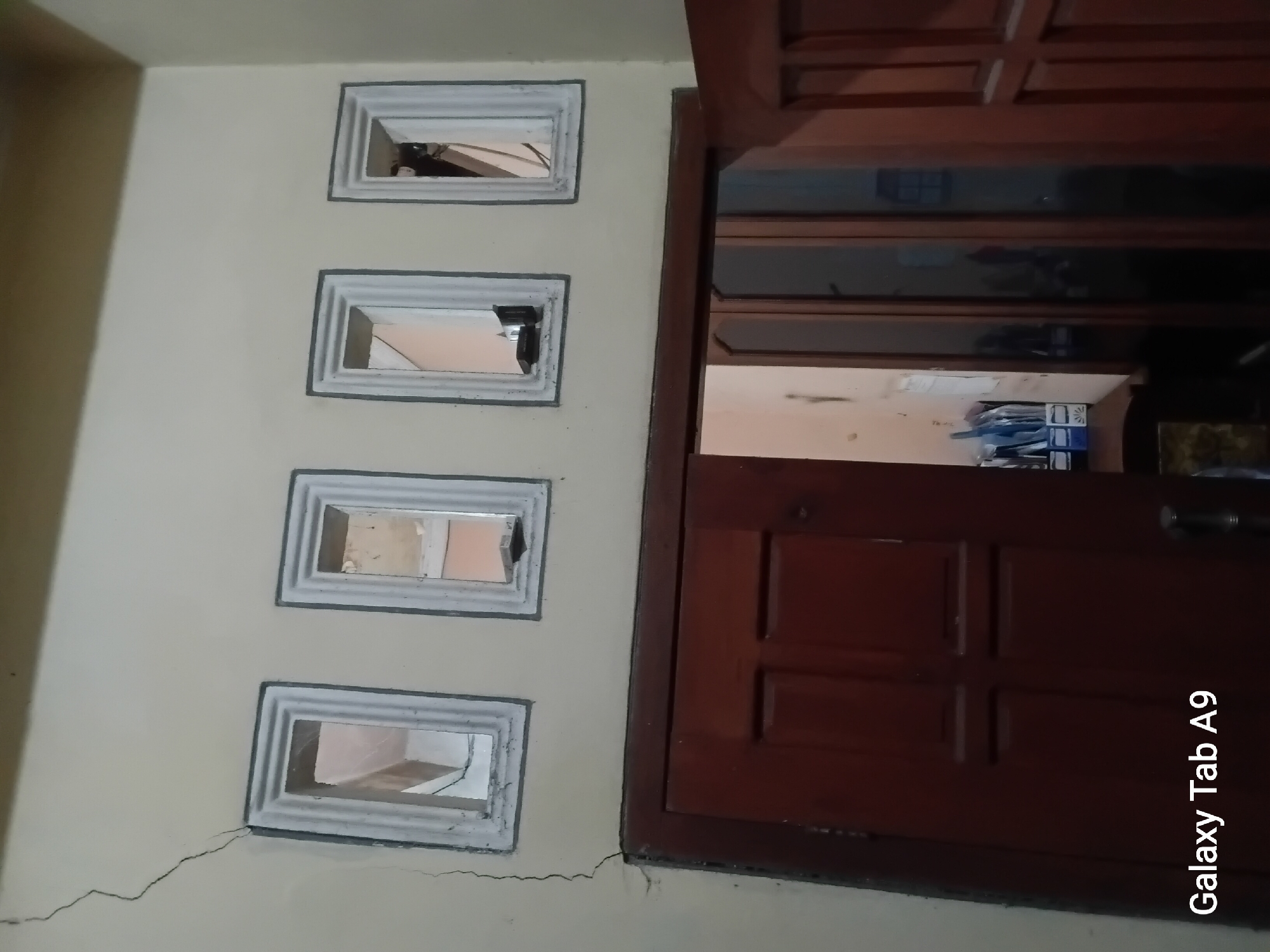 The image captures an interior view of a room, focusing on a section of a wall with four small rectangular openings and a dark brown wooden door below. The wall is painted a light yellow color and shows a visible crack extending from the top left corner downwards, intersecting with the upper frame of the leftmost opening. Each opening has a white, raised frame. Through the openings, glimpses of an interior space are visible, including what appears to be a room with some objects. The wooden door below is solid and has a panel design. To the left of the door, an opening reveals more of the interior, where a desk or surface holds a container with pens and other office supplies. The bottom left corner of the image contains a watermark displaying "Galaxy Tab A9". The scene suggests an indoor setting, possibly a home or an office space. The location is identified as Trenggalek, Indonesia.
