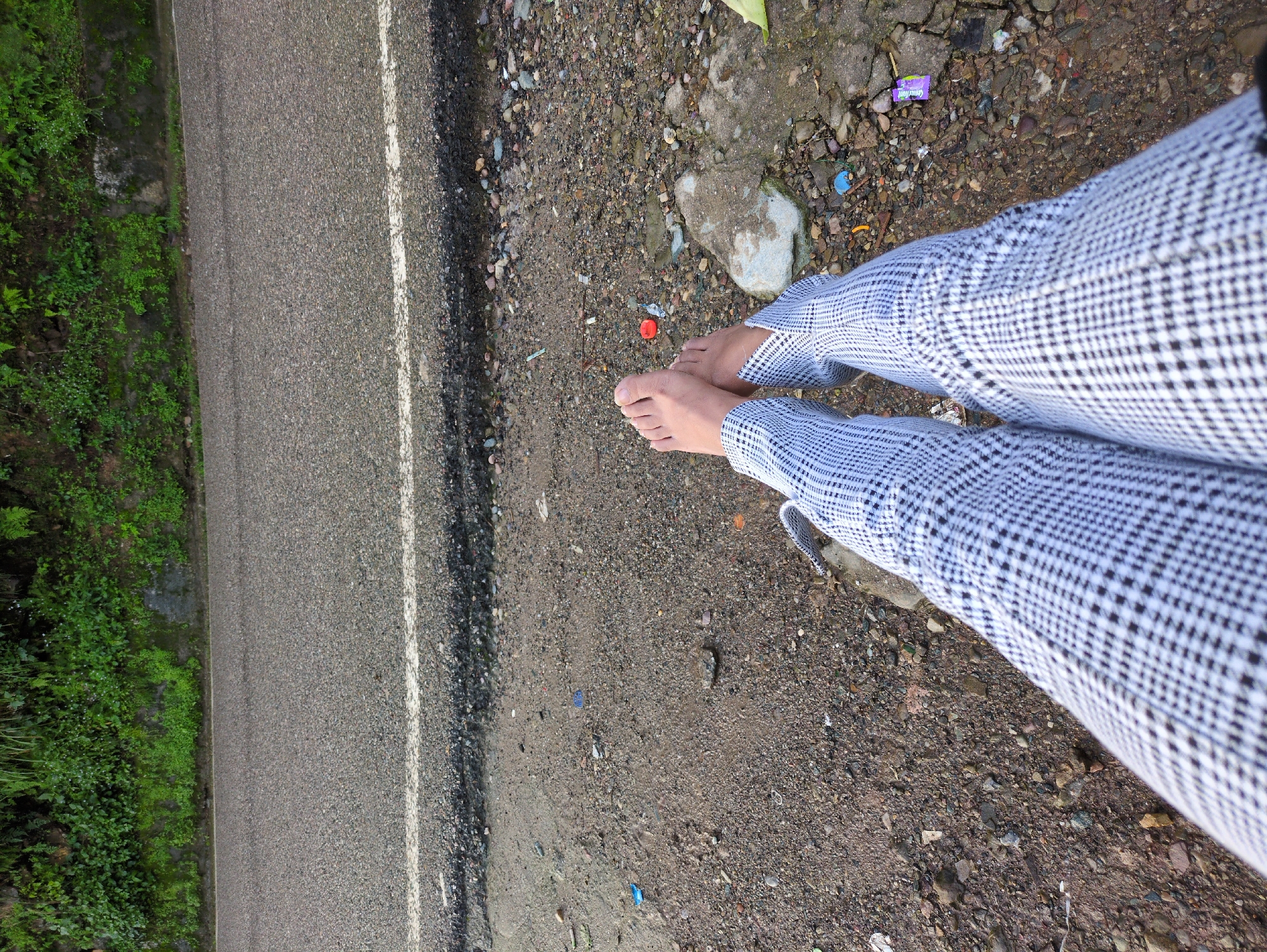 The image shows a person's bare feet and legs, wearing plaid pants, resting on the ground next to a road. The road appears to be an asphalt surface with a white line marking. The ground next to the road is covered with dirt, pebbles, and some scattered debris, including plastic wrappers. In the background, there is a lush green embankment with vegetation. The overall impression is that of a roadside scene in a natural environment, possibly near a hilly or mountainous area. While the image captures a specific moment, there are no clear indicators to pinpoint the exact location within Dharamshala, India, beyond it being a roadside setting.