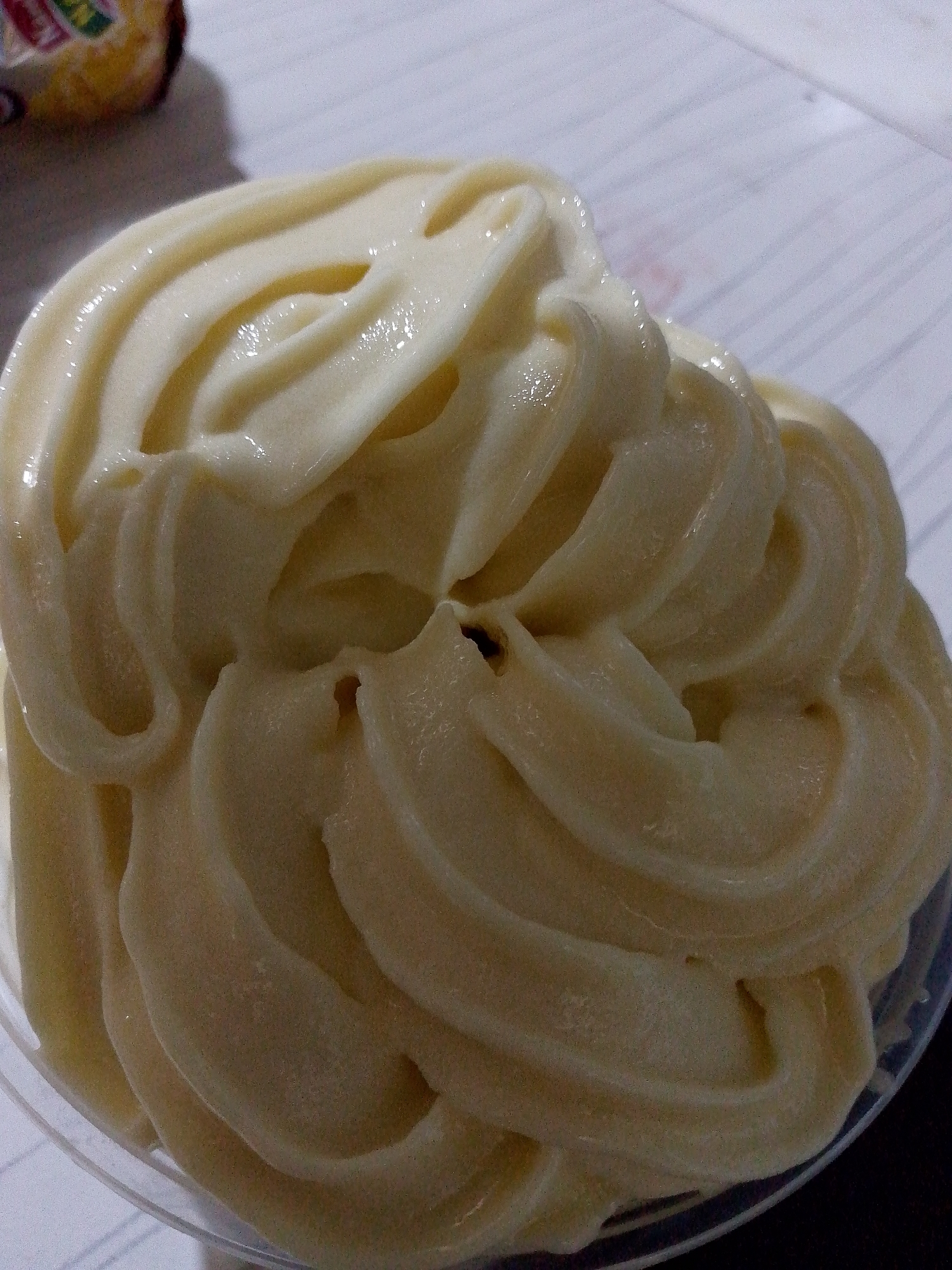 A close-up, overhead shot shows a swirl of pale yellow, soft-serve ice cream in a clear plastic cup, positioned on a white, lined surface. The ice cream has a glossy sheen and displays prominent ridges from the soft-serve machine. A portion of a colorful, circular object, possibly a wrapper or cookie, is visible in the upper left corner, out of focus. The background is a white surface with faint, parallel lines.