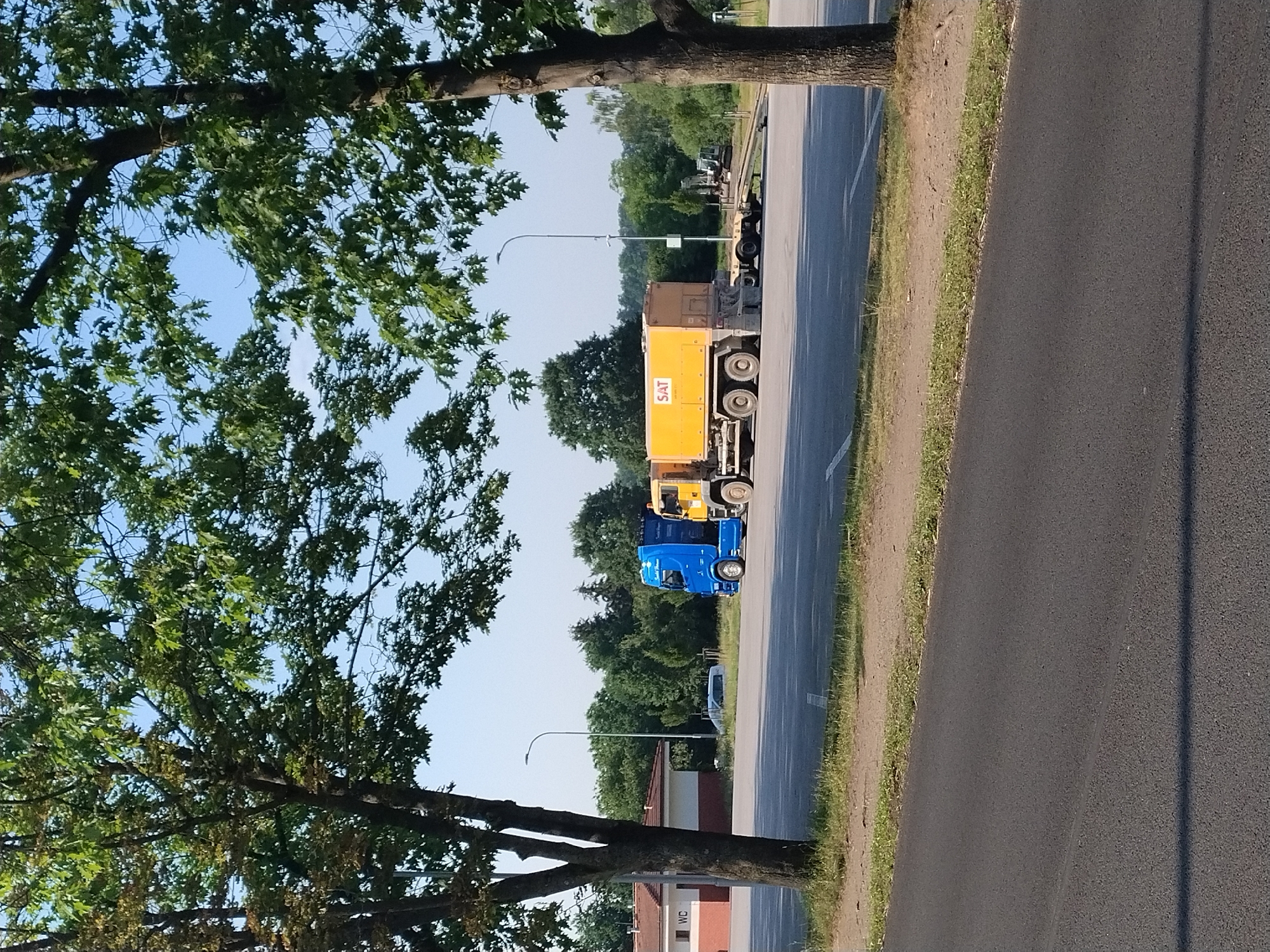 In Hluboká nad Vltavou, Czechia, on a clear day, two trucks are parked in an open lot. The truck on the left is blue, and the truck on the right is yellow with a large tank on its back that has "SAT" written on it. A building with a red roof and white walls, labeled "WC," is visible in the background. Trees with green leaves surround the area, and a paved road is in the foreground. The scene is calm and depicts a typical roadside rest area.