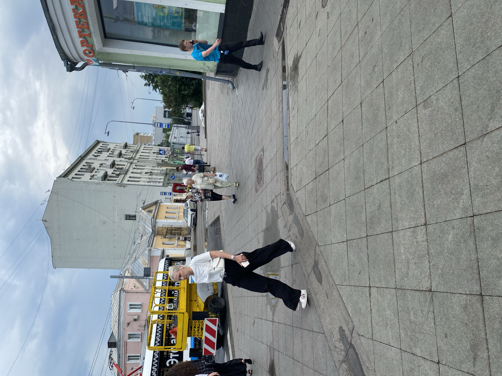 The image depicts a street scene in Moscow, Russia, on a cloudy day. A woman in black pants and a white t-shirt is walking across a grey tiled sidewalk, holding a smartphone. To her left, a yellow construction lift is parked on the street. In the background, there are buildings of various architectural styles, including a tall, modern white building and some older, pastel-colored structures. Several other pedestrians are visible on the sidewalk, and cars are present on the street. A shop with the word "ОРТЕКА" (likely meaning pharmacy) is visible on the right side of the image. The overall impression is of a bustling urban environment.