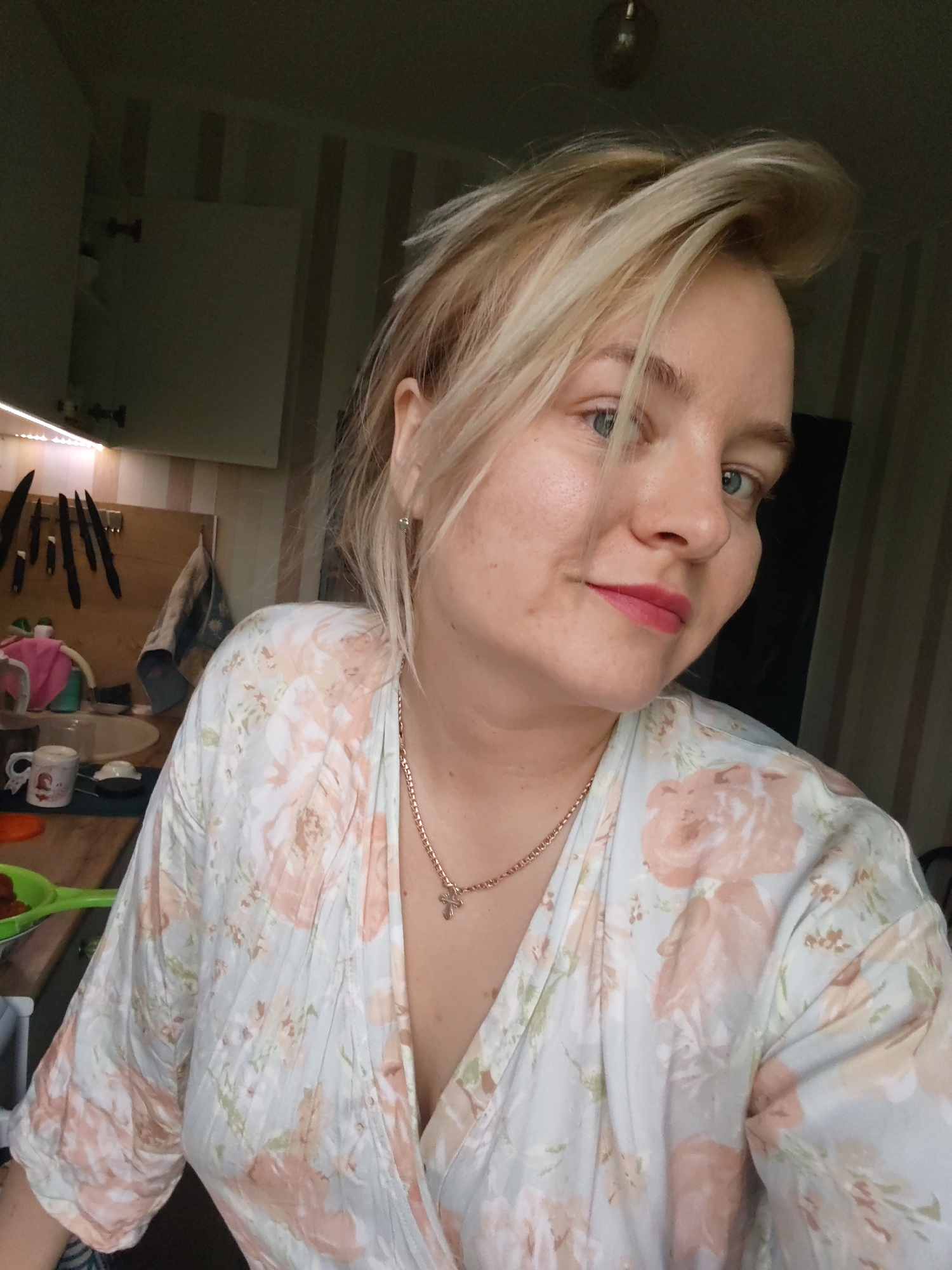 A medium shot captures a blonde woman with blue eyes, wearing a floral wrap dress and a gold necklace with a cross pendant. She has fair skin and is wearing pink lipstick. Her hair is styled in a tousled manner, with some strands falling across her forehead.
She appears to be in a kitchen setting. The background includes kitchen cabinets, utensils, and cooking items. A set of knives is mounted on a wooden surface. The walls are decorated with vertical striped wallpaper. The overall lighting is dim.
Based on the information provided, the location is inferred to be in a kitchen within the city of Izhevsk, Russia, although there is no explicit visual evidence to confirm the city.