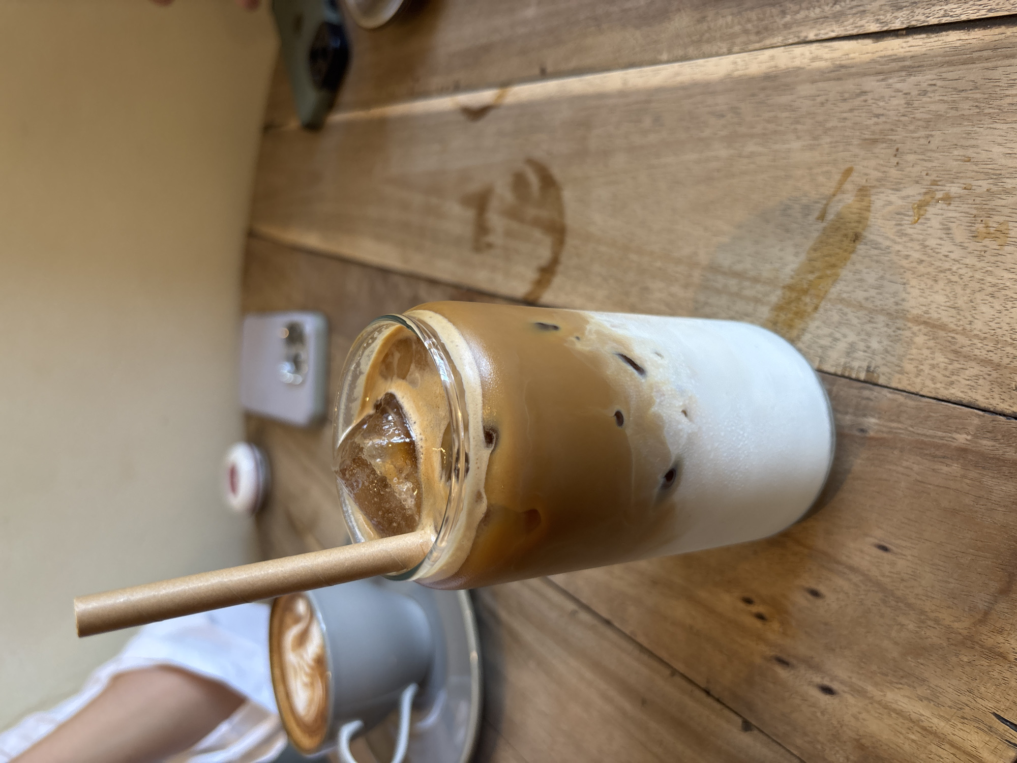 On a wooden table in Kuta Selatan, Indonesia, there is a glass of iced latte with a paper straw. Next to it, there is a cup of cappuccino and a cell phone. A person is visible wearing a white shirt.