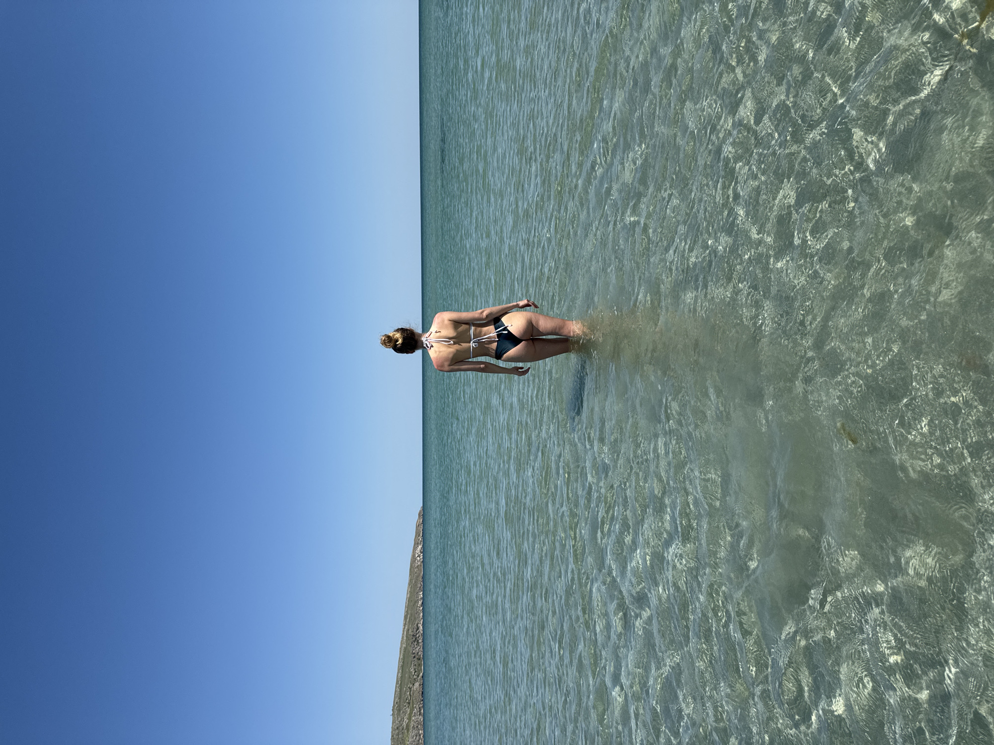 A woman is standing in shallow, clear water under a cloudless blue sky. She is facing away from the viewer, wearing a dark bikini with white straps. Her hair is tied up in a bun. The water appears to be clear and shallow, with ripples and variations in color. A land mass, possibly a cliff or shoreline, is visible on the left side of the frame. The location is Мысовское сельское поселение, Ukraine.