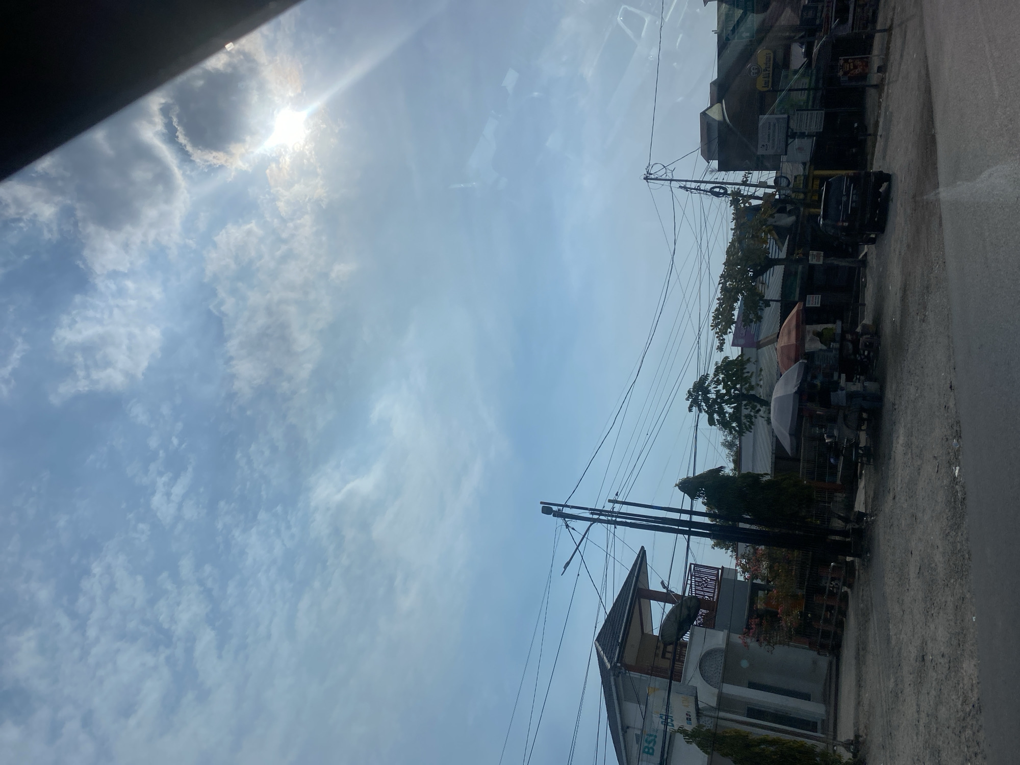 The image depicts a street scene in Gambut Barat, Indonesia, under a partly cloudy sky. The sun is visible, shining brightly through the clouds, casting a glare on the right side of the frame. A building with a sign that reads "BSI" is on the left, with a balcony and a satellite dish. Across the street, there are several small shops and stalls, some with umbrellas providing shade. A black SUV is parked in front of one of the shops. Utility poles with numerous wires crisscross the sky above the street. The ground is a mixture of dirt and asphalt. Two trees are visible on the left side of the road, and a few more trees and foliage are scattered throughout the scene. The overall impression is of a typical commercial area in a town or city.