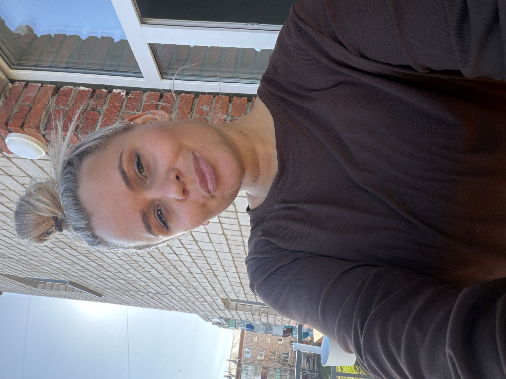 A selfie-style image captures a young adult woman from an overhead perspective, with her head tilted to the right. She has light-colored hair, likely blonde or greyish blonde, pulled back into a bun with a dark hair tie. Her eyes are open, with visible eyeliner and mascara, and her lips are slightly parted. Her left ear is prominent, showcasing multiple piercings including studs on the lobe, helix, tragus, and conch. She wears a dark brown, long-sleeved top.
The background is a mix of architectural elements and open sky, suggesting an outdoor setting, possibly a balcony or elevated area in Sarapul, Russia. Immediately behind and to the left of her head is a red brick wall with a white, circular light fixture. Above this, a white window frame reflects a distant building or the sky. Below and to the left of her face, a light-colored, square-patterned tiled surface is visible, extending towards the horizon. In the distance, under a bright, sunny, light blue sky, several residential or urban buildings with numerous windows can be seen, along with a bare tree. A white cup with a lid and straw is partially visible in the lower right corner, suggesting she is enjoying a beverage. The bright sunlight indicates it is daytime with clear weather. The woman's expression appears relaxed or neutral. No text is visible in the image.