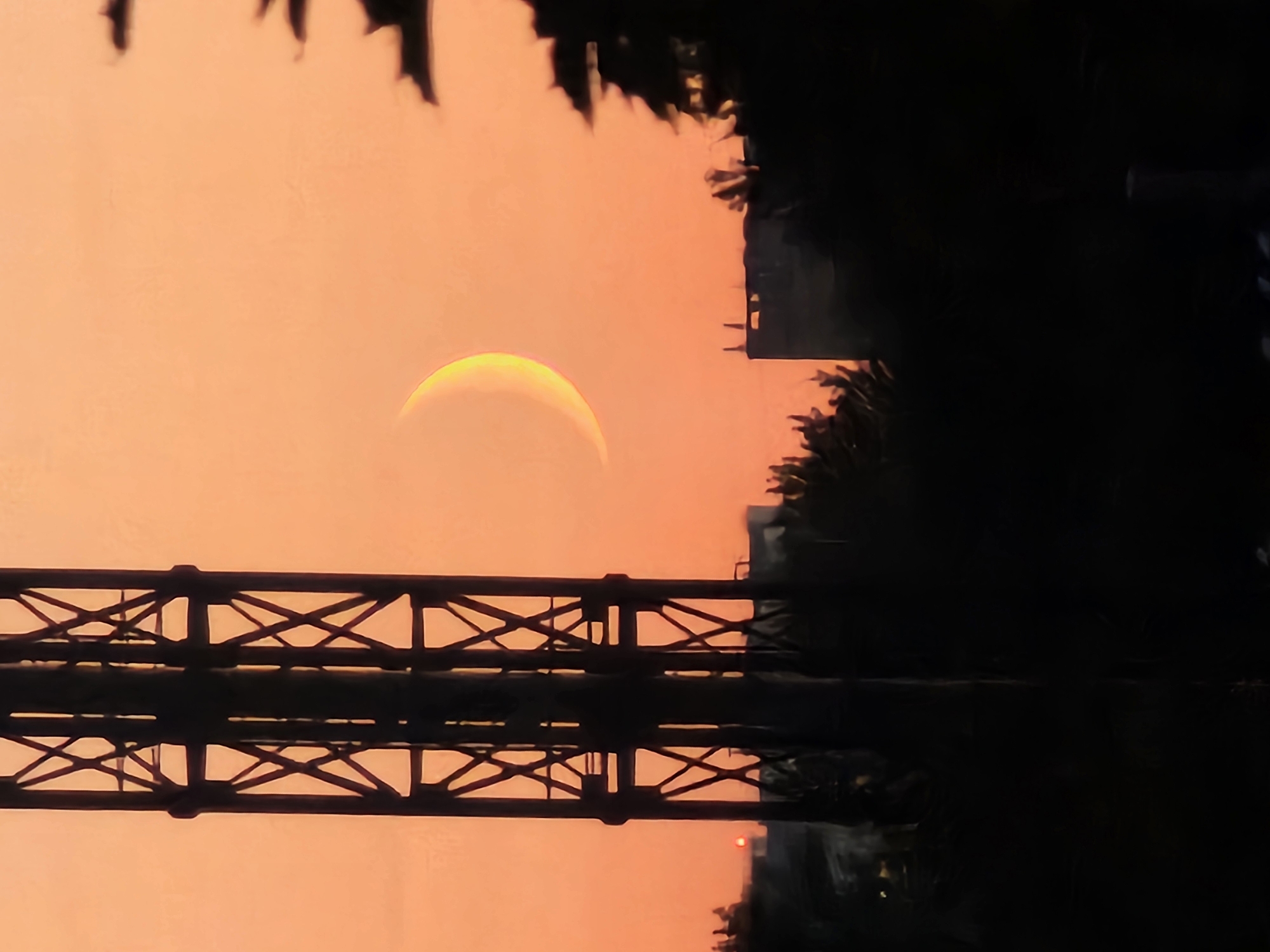 A crescent moon is visible in the orange sky over Khimki, Russia. In the foreground, a dark silhouette of a metal tower is on the left, and trees and buildings are silhouetted against the sky on the right. The scene appears to be a cityscape at dusk or dawn.