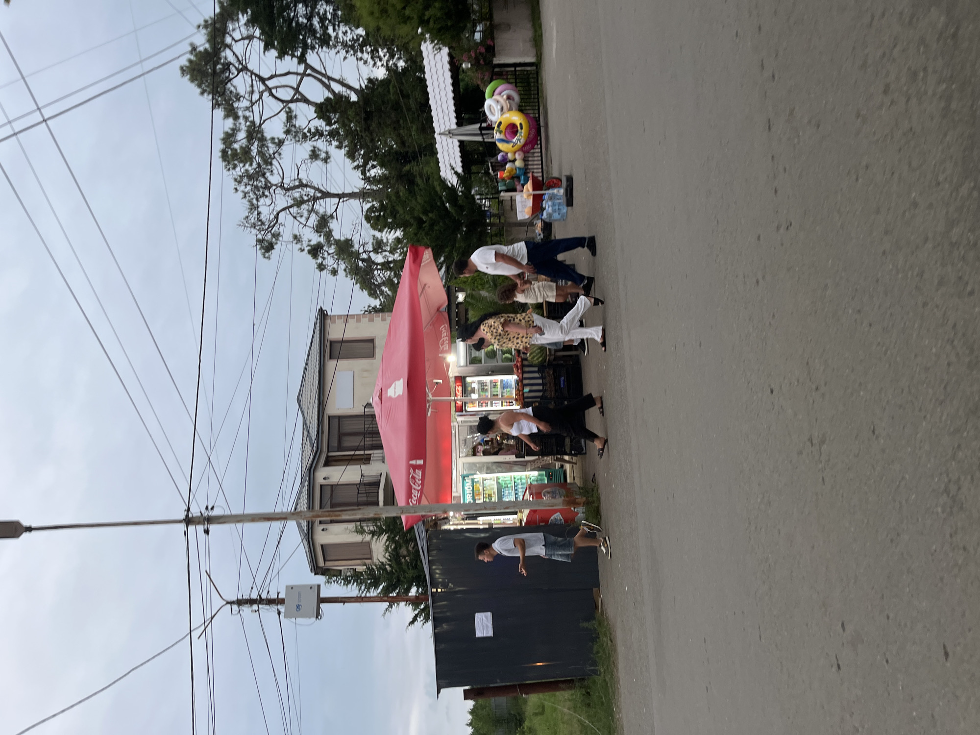 This image captures a street scene in Ureki, Georgia, on an overcast day. A small convenience store with a bright red umbrella covers its storefront, displaying various drinks and possibly snacks inside. Several people are visible. A young man in a grey polo shirt and shorts walks past a dark grey metal structure on the left. A woman in a white top and black pants walks towards the store. Further down, a family is gathered near a collection of colorful inflatable rings and a cart with bottles of water, suggesting a recreational area or merchandise for sale. In the background, a multi-story building with balconies stands behind the store, and tall trees with dense foliage frame the scene. Overhead, a complex network of electrical wires crisscrosses the sky, supported by utility poles. The ground is paved with asphalt, showing signs of wear.