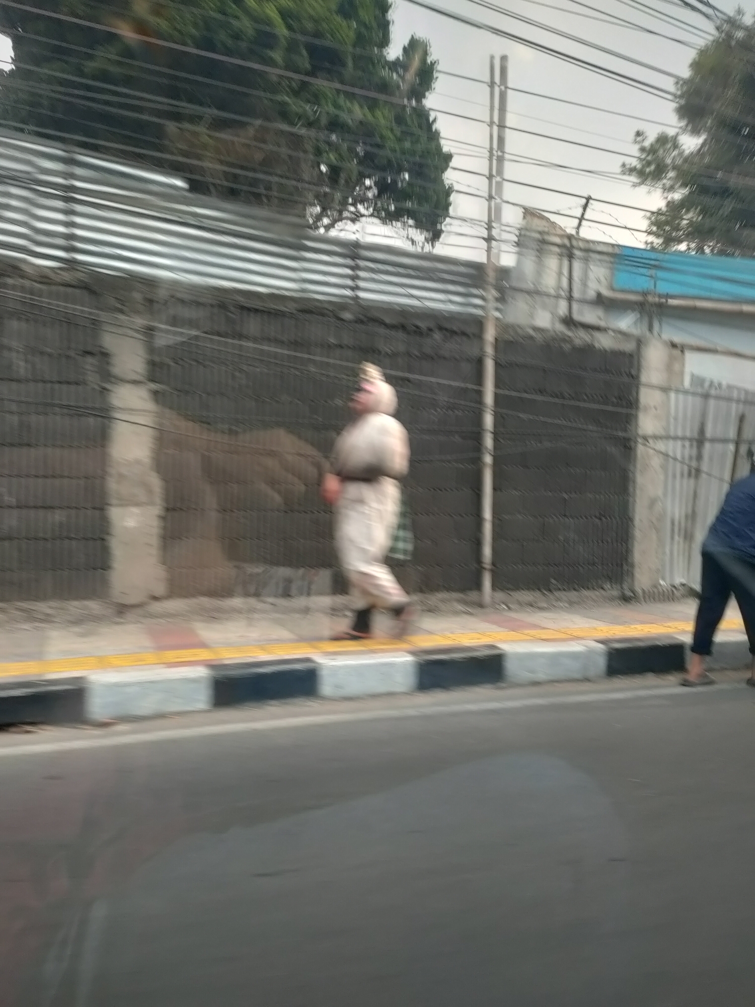 The image, captured from a moving vehicle, shows a street scene in Lembang, Indonesia. A person wearing a light-colored, possibly traditional, outfit with a head covering is walking on a sidewalk next to a textured dark wall. The sidewalk has yellow tactile paving and a border of alternating grey and black concrete blocks. Behind the wall, there are power lines and trees, with a corrugated metal fence visible in the background. Another person, wearing a dark blue top and dark pants, is also on the sidewalk further to the right, with their feet visible at the edge of the frame. The road in the foreground is asphalt.