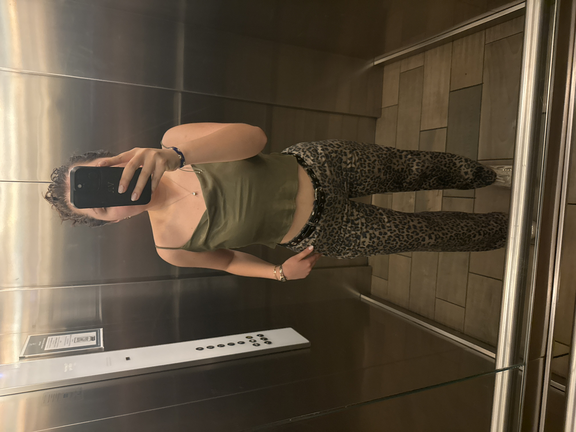 A woman is taking a selfie in an elevator. She has curly hair and is wearing a green tank top and leopard-print pants with a black belt. She is holding a black phone. The elevator has stainless steel walls and a control panel. The floor appears to be tiled. The location is not specified or identifiable within the image, and Mexico City is not discernible.