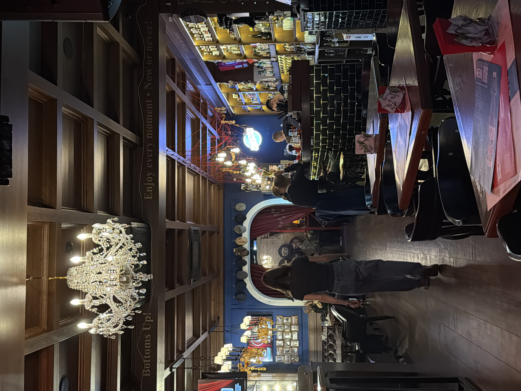 The image shows the interior of a restaurant or bar, likely in Moscow, Russia. The scene is a long, narrow room with a high ceiling featuring exposed wooden beams and an elaborate chandelier. The walls are adorned with framed photographs and memorabilia, suggesting a historical theme. Several people are present, including patrons seated at tables and bartenders behind the bar. Tables are set with menus and what looks like promotional material. The bar is well-stocked with bottles, hinting at a wide selection of alcoholic beverages. There's a sign with the phrase "Bottoms Up!" and "Enjoy every moment. Now or never!" implying a lively atmosphere. The lighting is warm, creating a welcoming ambiance. The image appears to be taken at a time when the place is fairly occupied with patrons.