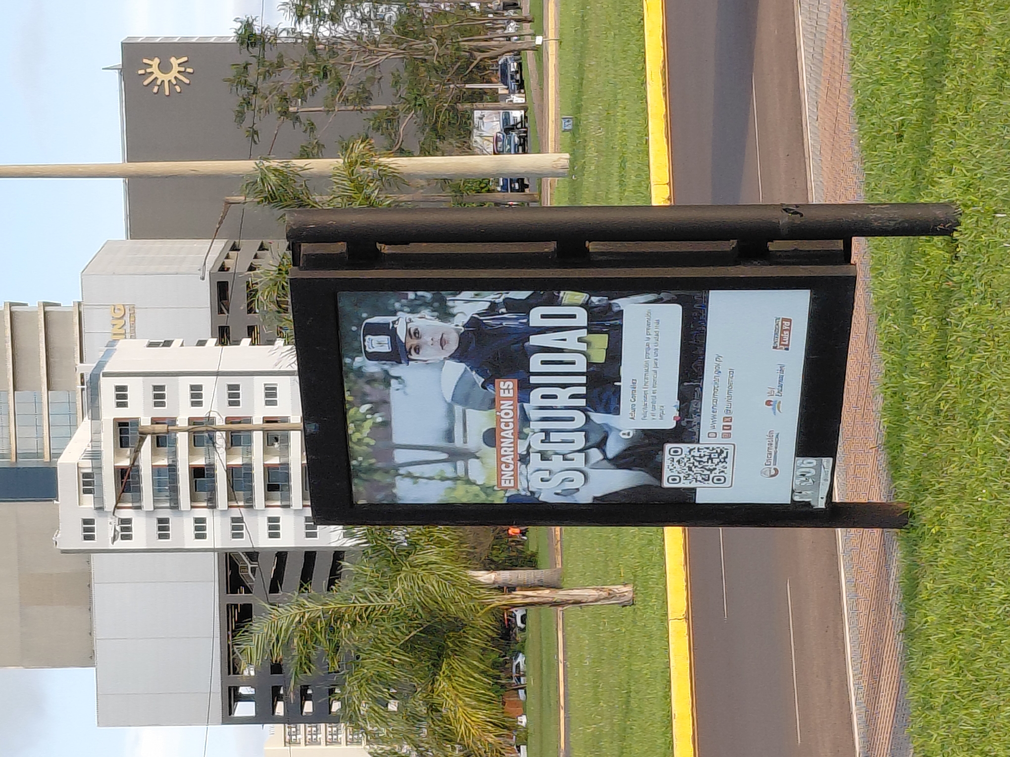A vertical medium shot captures an outdoor digital billboard displaying a public service announcement against a backdrop of modern city architecture and greenery. The billboard's content features a woman in a security uniform and the text "ENCARNACIÓN ES SEGURIDAD" (Encarnación is Security). Below this, smaller text describes the importance of prevention for a safe city. A QR code and social media handles are also visible, along with logos for "Encarnación" and "Intendente Luis Yd."
In the background, several tall buildings rise against a hazy sky, including a white building with multiple balconies and a darker structure with a distinctive sun-like logo. A palm tree is prominently positioned to the left of the billboard, and other trees and manicured green grass surround the display. A street with yellow and white markings is visible in the lower portion of the image, with a few cars parked or driving in the distance. The scene appears to be a public urban space, likely a plaza or promenade, in Encarnación, Paraguay, given the text on the billboard. The overall atmosphere is bright and daytime, with overcast skies.