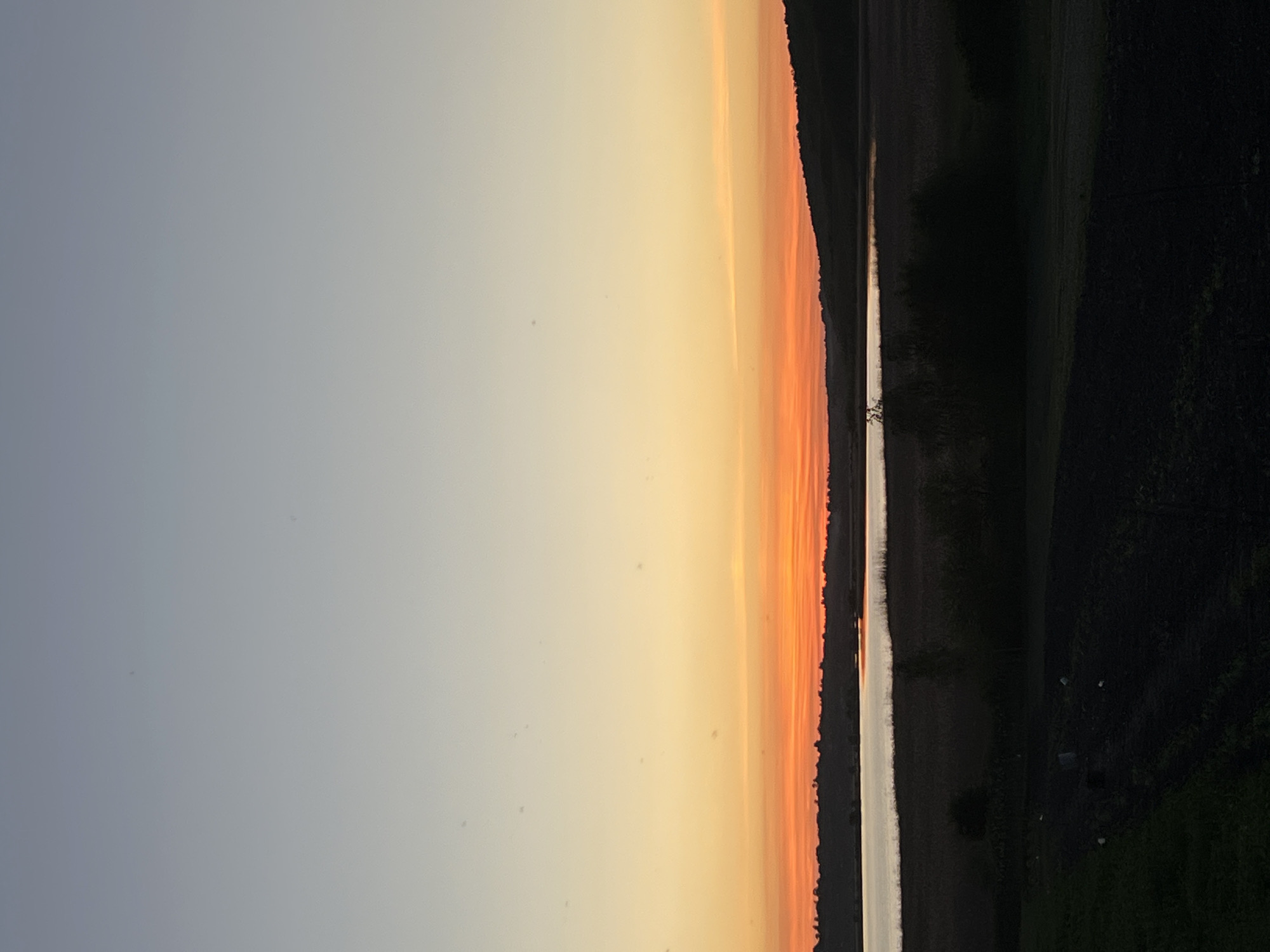 The image presents a landscape scene at dusk, likely in a rural area near Vertelka, Ukraine. To the left, the sky transitions from a light gray-blue to a gradient of orange, yellow, and red, indicating a sunset. A body of water, possibly a lake or river, reflects some of these colors in its center. The right side of the image shows dark vegetation and terrain, suggesting a forest or overgrown fields. The overall atmosphere is one of natural beauty at twilight.