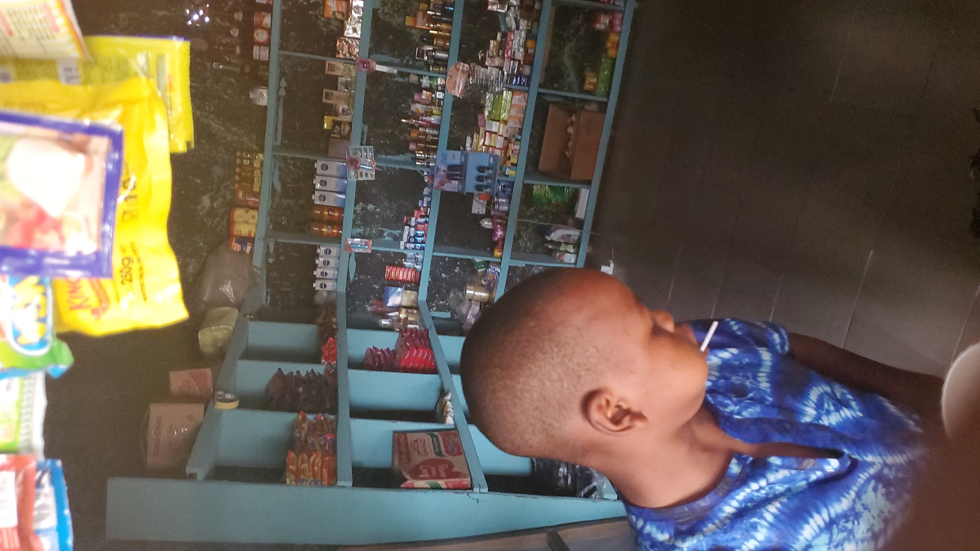 A young boy with a short haircut, wearing a blue tie-dye shirt, is the primary subject in this image. He is seen from the side, with his head tilted back and a white lollipop sticking out of his mouth. The boy is indoors, in what appears to be a small shop or market stall.
The background is filled with shelves stacked with various products. These include small boxes, packets, and bottles of what could be food items, toiletries, or other consumables. The shelves are painted a light blue, and the wall behind them has a patterned wallpaper. To the left of the shelves, there are some hanging yellow and purple plastic packages, likely containing snacks or other goods. The flooring is a dark, tiled surface.
The lighting suggests it is daytime, possibly late afternoon, given the warm tones and shadows. There is no indication of weather conditions. The setting feels like a local marketplace or a small convenience store in Jalingo, Nigeria. The boy's posture and the lollipop suggest he is enjoying a treat, possibly while accompanying someone shopping or simply browsing. The overall mood is casual and everyday.
There is some visible text on the packaging, including "250g" on a yellow bag, and the word "KING" is partially visible on another package. The image's orientation is slightly skewed, making it appear rotated.