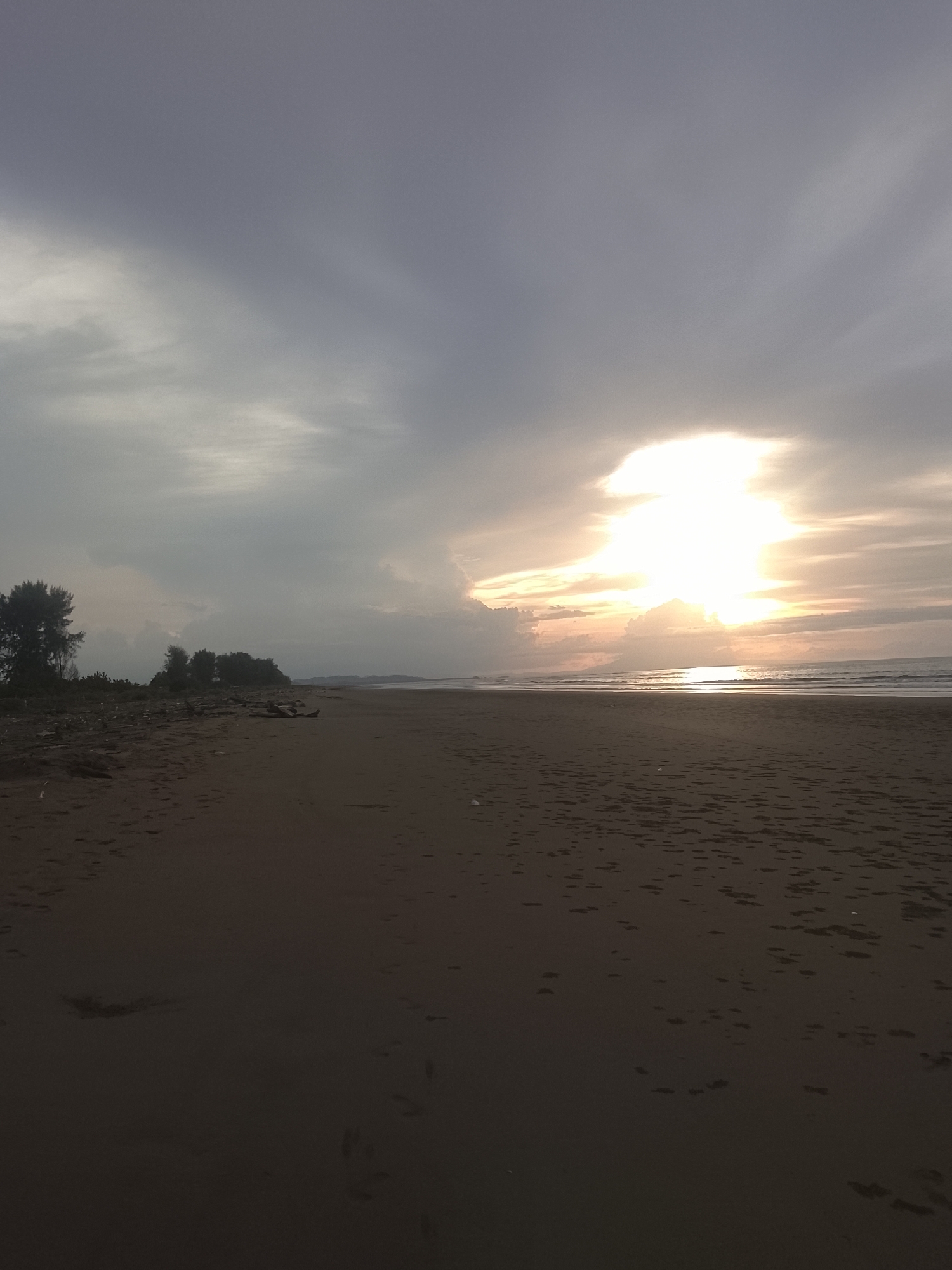The image depicts a beach scene at dusk. The sky is overcast with a bright, diffused sun peeking through the clouds on the right side of the frame, casting a warm glow over the ocean. The ocean itself is calm with gentle waves lapping at the shore. The beach is composed of dark sand, with some debris scattered along the tideline, including driftwood. In the background, a line of trees and vegetation forms a natural barrier along the coast. The overall impression is one of a tranquil evening by the sea. This scene could be located in Reudeuep, Indonesia, known for its coastal beauty.