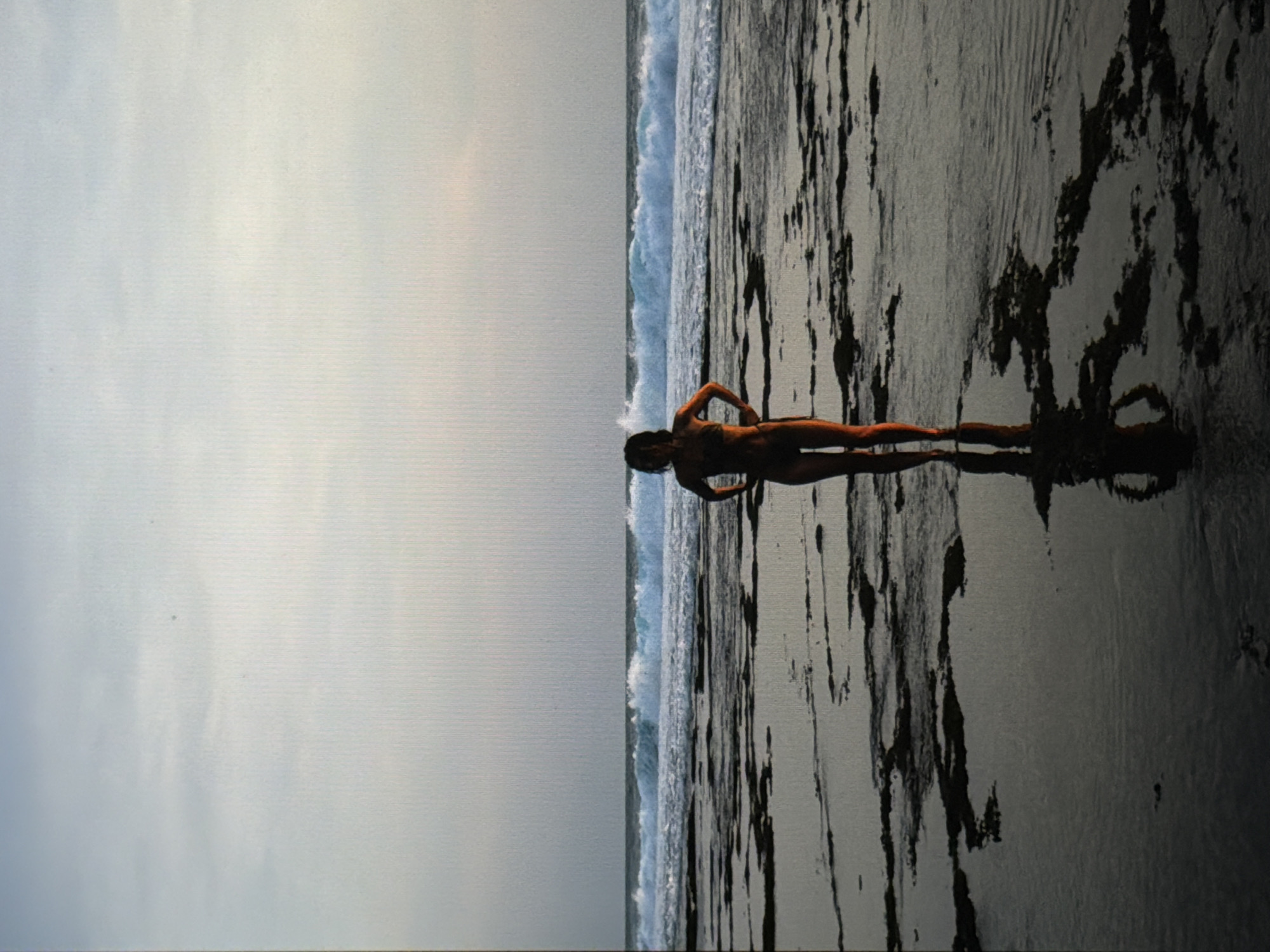 A woman stands on a black sand beach in North Kuta, Indonesia, near the ocean waves. The sky is overcast.