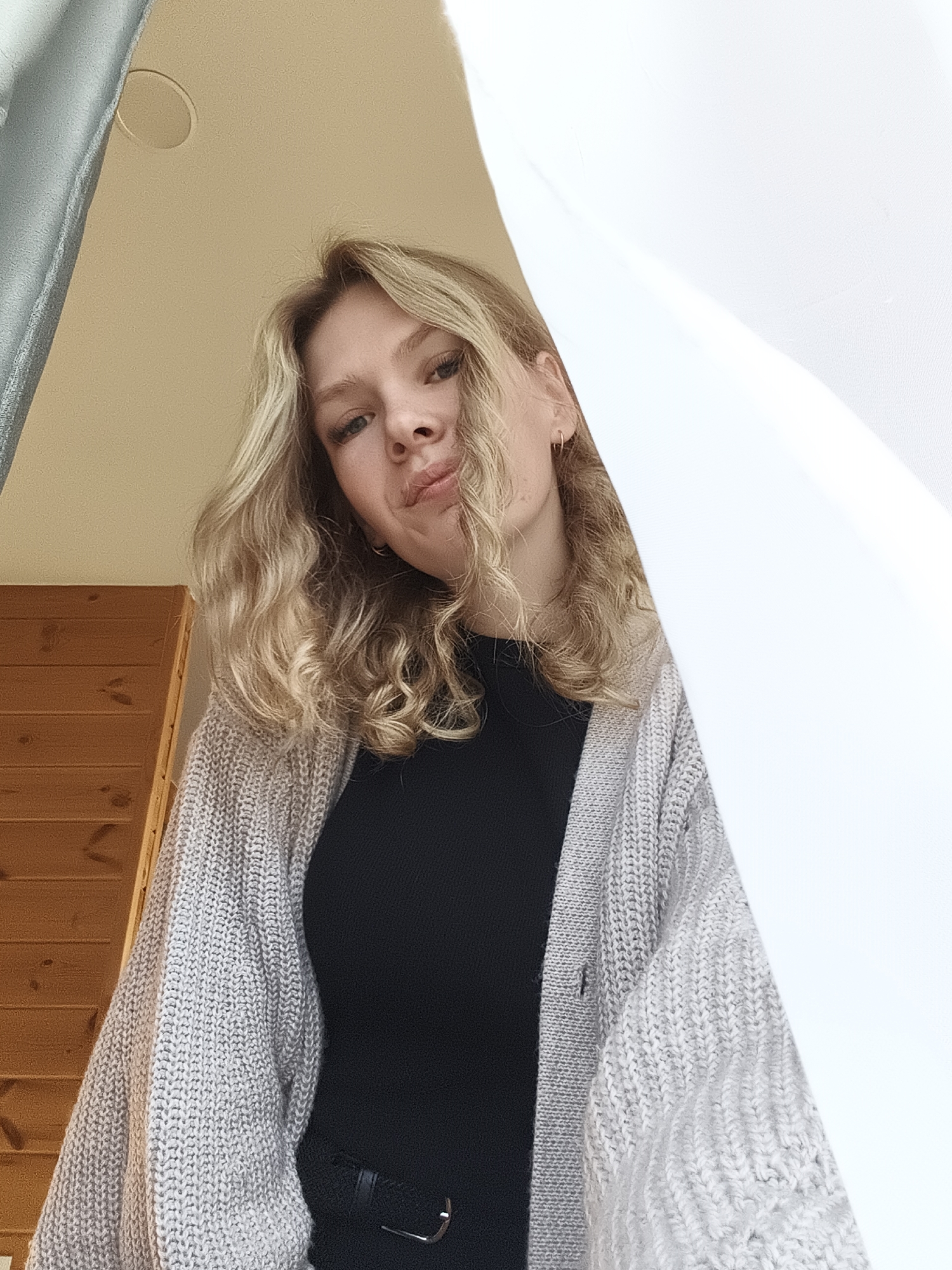A young woman with blonde, curly hair is looking down at the camera with a slight pout. She is wearing a black top and a light gray, knitted cardigan. Part of her is framed by a white curtain or sheet to her right and a gray one to her left. In the background to her left is wooden wall paneling. The location is ambiguous; there is nothing to indicate that this image was taken in the city of Русский Вожой, Russia.