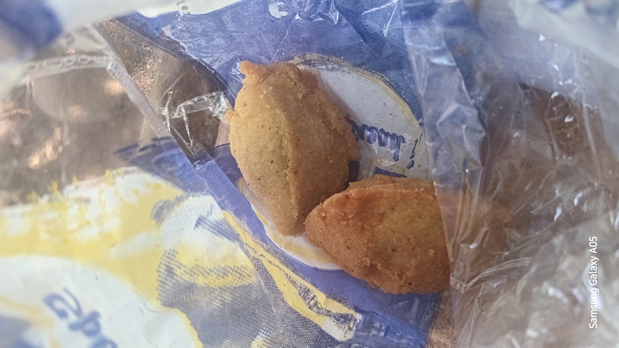 Two fried food items, possibly chicken nuggets or croquettes, are nestled inside a crinkled plastic bag with blue and yellow patterned paper lining. The food is golden brown and appears to have a textured, crispy coating. The background shows the blurry interior of the bag and some of the paper's design, which includes what looks like text or logos. The image has a watermark in the bottom left corner that reads "Samsung Galaxy A05". The lighting is uneven, casting shadows and highlights on the food and the bag. No people or external environment are visible. The scene is indoors, likely a food packaging.