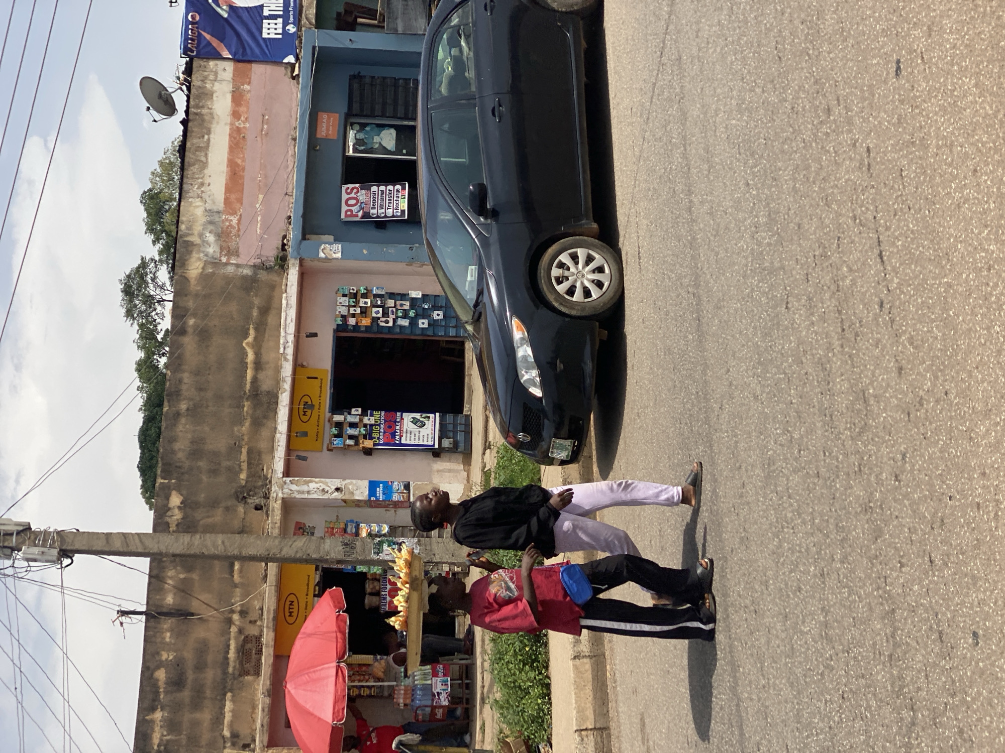 This image depicts a street scene in Jos, Nigeria, under a bright, sunny sky. Two young people are the primary subjects, with one standing and looking up, wearing a black hoodie and light purple pants, and the other bent over slightly, wearing a red t-shirt and black pants with white stripes. The second person is holding a tray of what appears to be corn on their head.
They are standing on a paved road next to a row of shops. A black car is parked on the side of the road. The shops have various signs, including "MTN," "JUMIAO," and a "POS" sign advertising deposit, withdrawal, and transfer services. There are also signs for "LALIGA" and "FEEL THE Sports Premi." A red umbrella is partially visible on the left side of the image, likely belonging to a vendor. Power lines and a concrete utility pole are prominent in the background. The overall scene suggests a busy commercial area, likely during daylight hours given the strong shadows. The people's posture and expressions are neutral, with the person looking up seeming to be in thought or observing something.