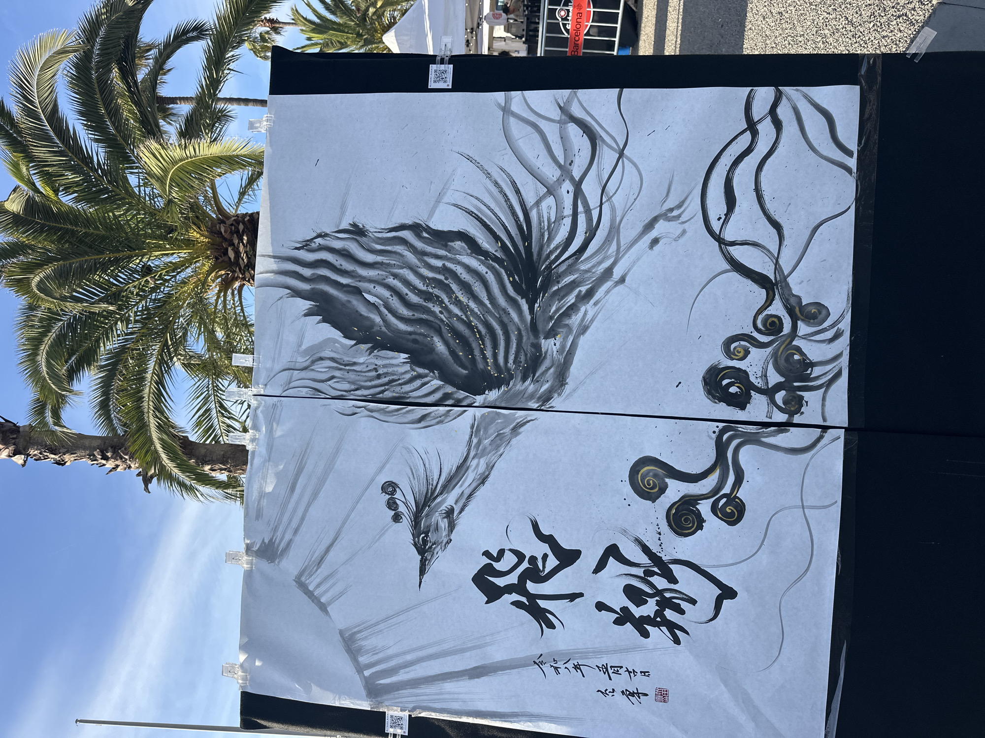 The image shows two pieces of East Asian-style art displayed outdoors, possibly at a market or art fair. The artwork features a stylized bird with flowing lines and traditional calligraphy, rendered in black ink with gold accents on a light background. The two panels are joined together and mounted on a dark display board. The sky is visible, with palm trees providing a backdrop, suggesting a warm, sunny climate. A partially obscured sign saying "Barcelona" is also visible. The location is Barcelona, Spain.