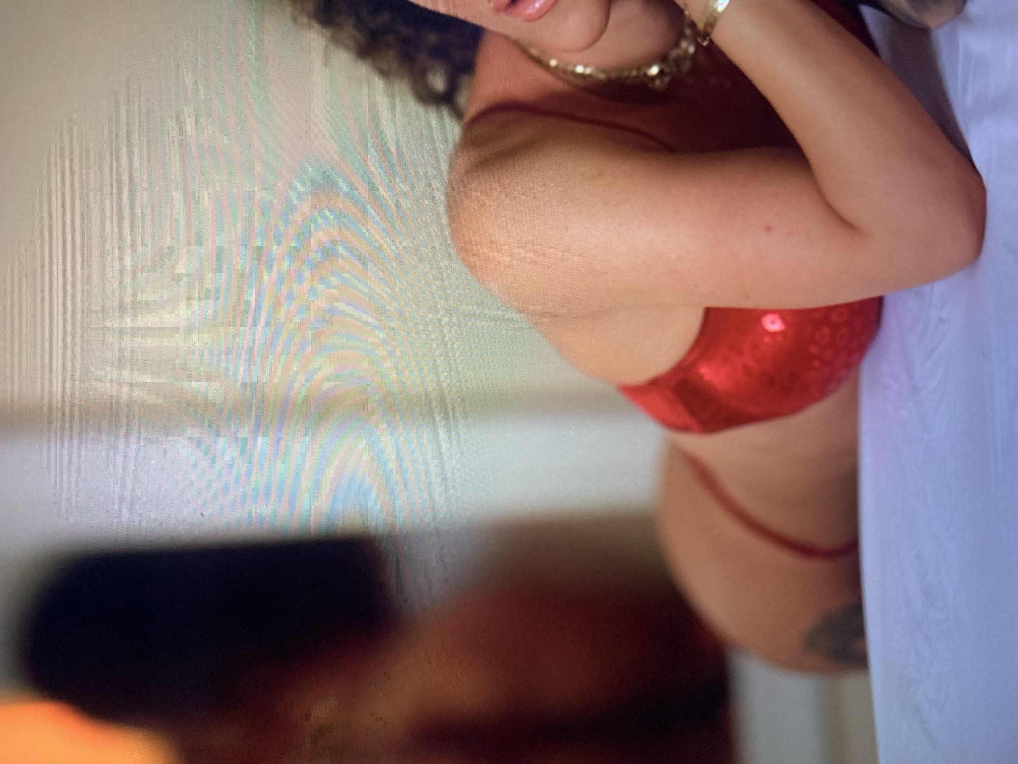 A person is lying on a white surface, wearing a red swimsuit and a gold necklace. The person's back and side are visible, with their head turned away from the camera. The background is blurred and appears to be a room with a light-colored wall.