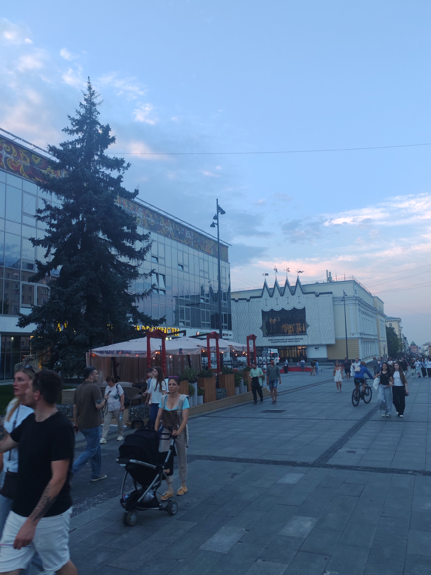 The image depicts a street scene in Nizhny Novgorod, Russia, likely in the evening. The foreground features a paved pedestrian walkway with several people walking, including a woman pushing a stroller. To the left, a large, modern building with reflective windows is visible, with a tall evergreen tree in front of it. A section of this building appears to be a cafe or restaurant with outdoor seating and umbrellas. Further down the street, another building with a distinctive architectural facade, featuring a jagged roofline and a large window displaying an interior, is present. Numerous people are walking along the sidewalk on the right side of the street. The sky is a light blue with some clouds, indicating the late afternoon or early evening. The overall atmosphere suggests a public urban space with ongoing activity.