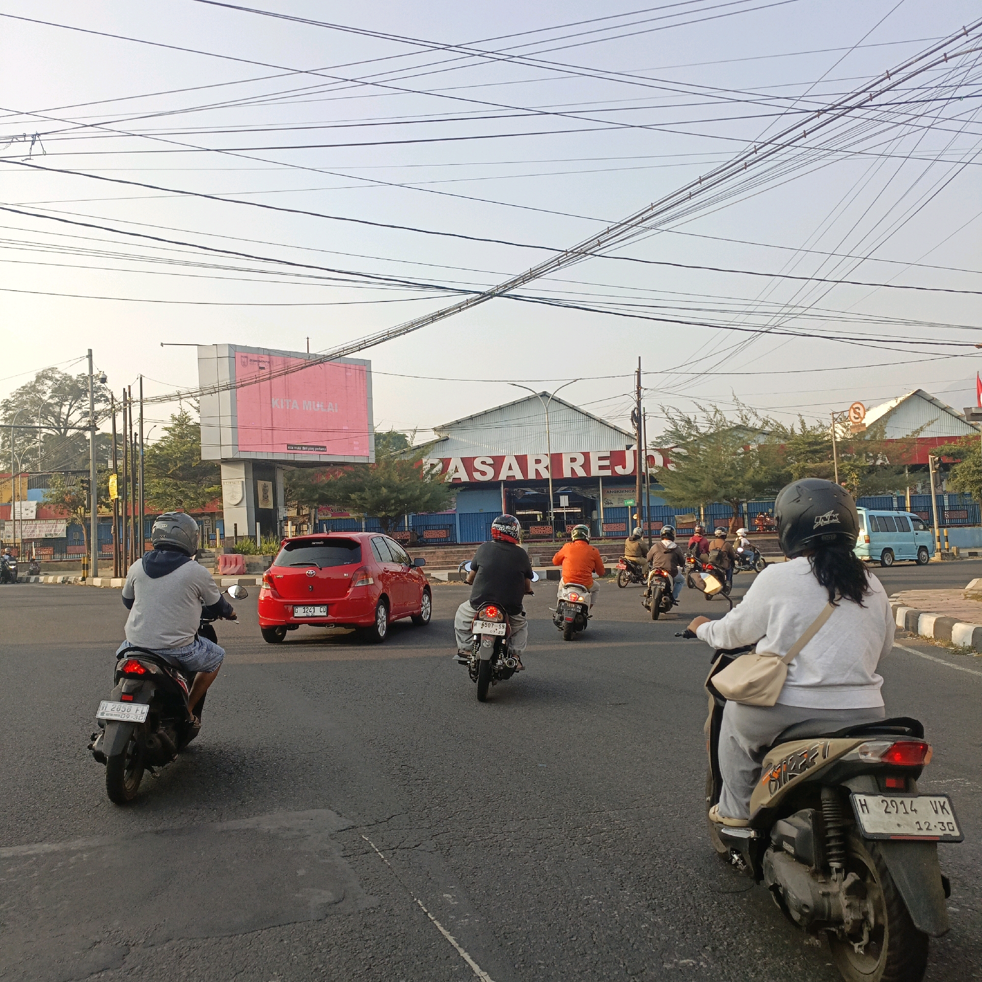 A street scene in Salatiga, Indonesia, features a bright, overcast sky with a complex network of overhead electrical wires crisscrossing the frame. In the mid-ground, a large, red billboard with white text reading "KITA MULAI" is prominently displayed above a government building. To the right of the billboard is a market building with red lettering that spells out "PASAR REJO". The street is paved with asphalt and marked with white lines. A variety of motorbikes and one red car are present on the road. Several individuals are riding motorbikes, some wearing helmets. In the foreground, a person wearing a gray hoodie and denim shorts rides a black motorbike. To their right, a red car with license plate G 1241 CD is driving. Further ahead, other motorbikes are visible, including one ridden by a person in an orange shirt. A light blue van is parked on the right side of the road. The environment appears to be an urban setting with some trees and a fence visible in the background.