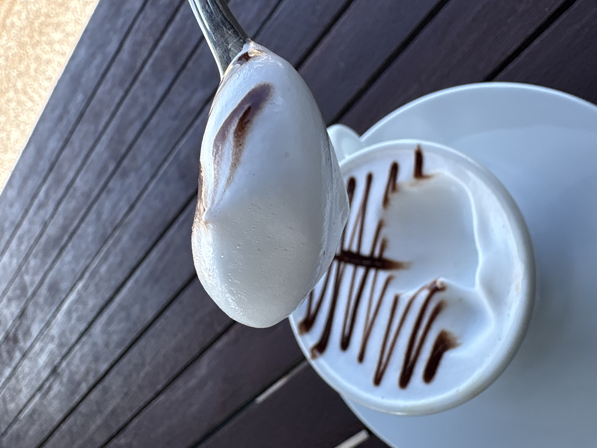 A high-angle, close-up shot shows a spoonful of meringue held above a white cup of a coffee beverage decorated with chocolate sauce. The cup sits on a white saucer. The background features a dark brown, horizontally-slatted surface resembling a wooden table. The location is likely a cafe in Bar, Montenegro based on the image context. No people are visible.