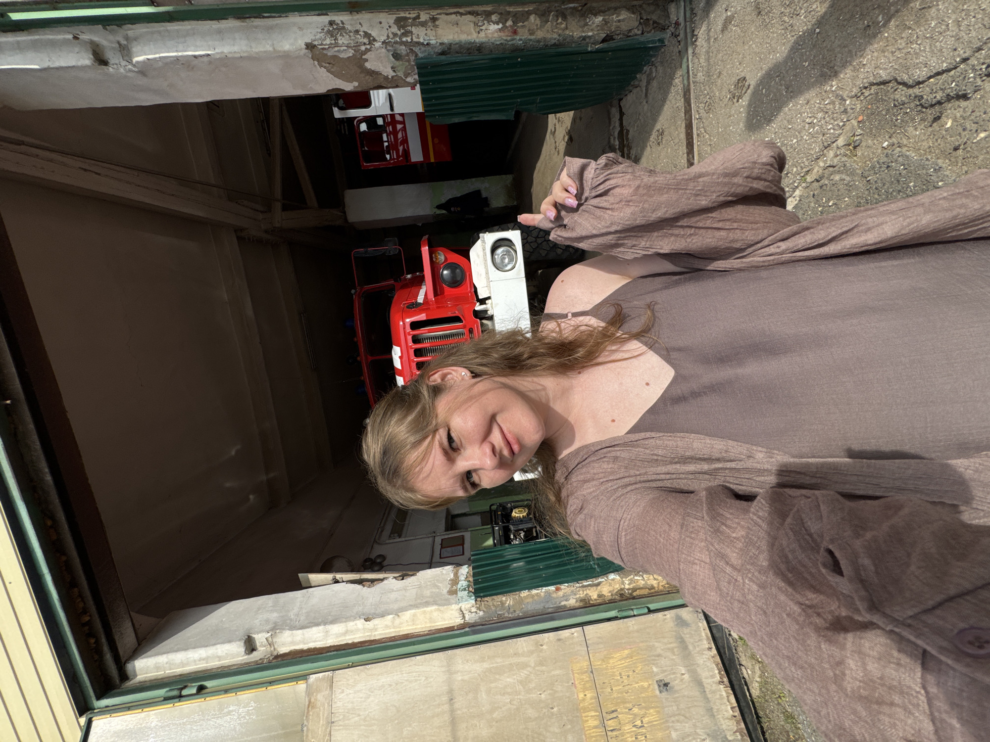 A woman with blonde hair, wearing a brown dress and a loose, brown, gauze-like cardigan, poses for a selfie in front of a fire station in Vereshchagino, Russia. She is smiling and giving a peace sign with her right hand. In the background, two red fire trucks are parked inside the open bays of the station. The fire station appears to be an older, rustic building with peeling paint on the concrete pillars. The ground in front of the station is paved with gravel and concrete.