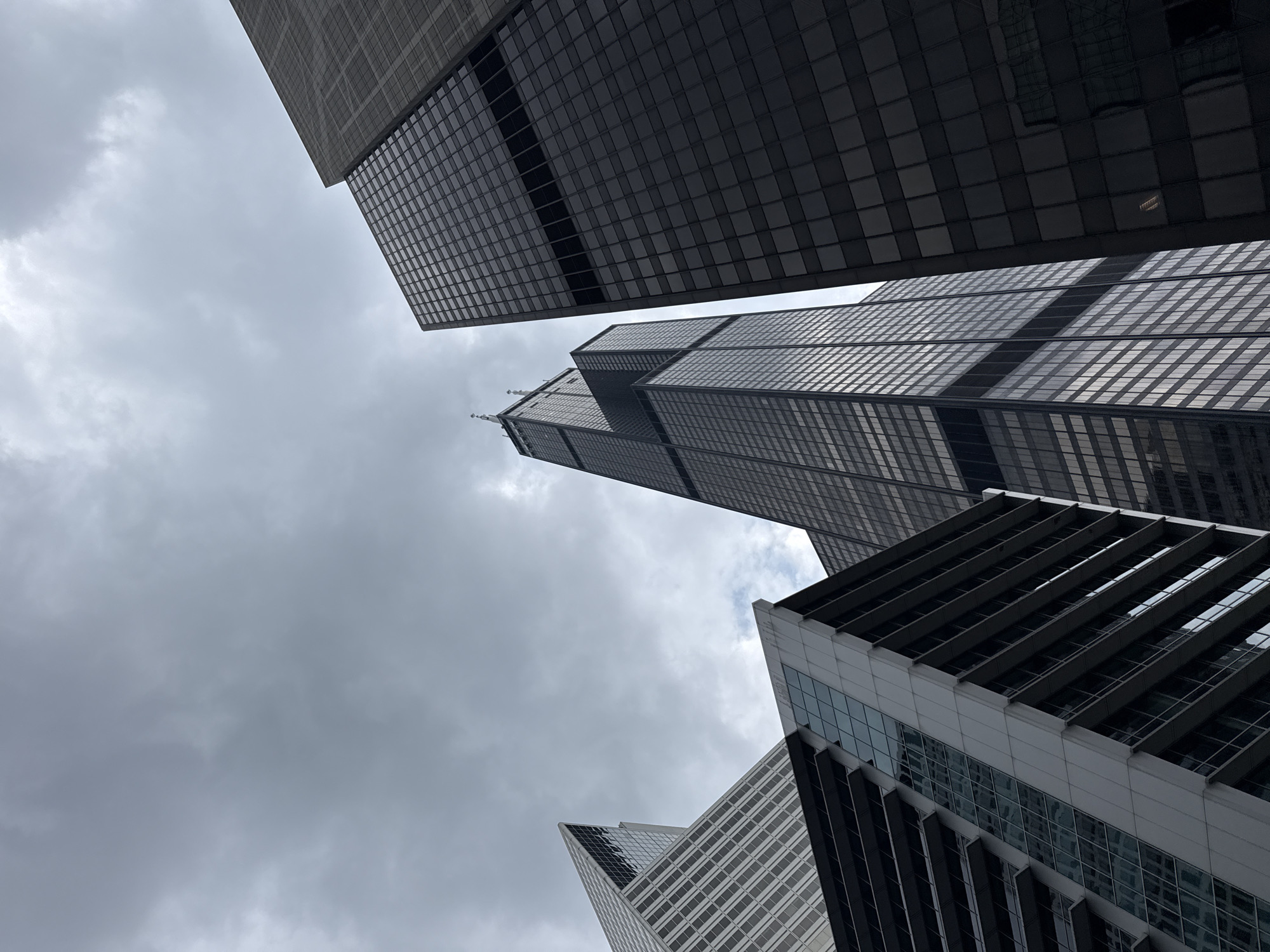 The photograph captures a low-angle view of several modern skyscrapers in Chicago, Illinois, USA, under a cloudy sky. The most prominent building is the Willis Tower (formerly Sears Tower), recognizable by its distinctive stepped design and twin antennas at the top. To its right, a darker, rectangular skyscraper with a grid-like pattern of windows dominates the frame. In the foreground on the left, a lighter-colored building with balconies and reflective windows is partially visible. The buildings are made of glass and metal, reflecting the overcast sky and the surrounding urban environment. The composition emphasizes the height and architectural styles of the buildings, creating a sense of scale and urban density. No people or specific actions are depicted.