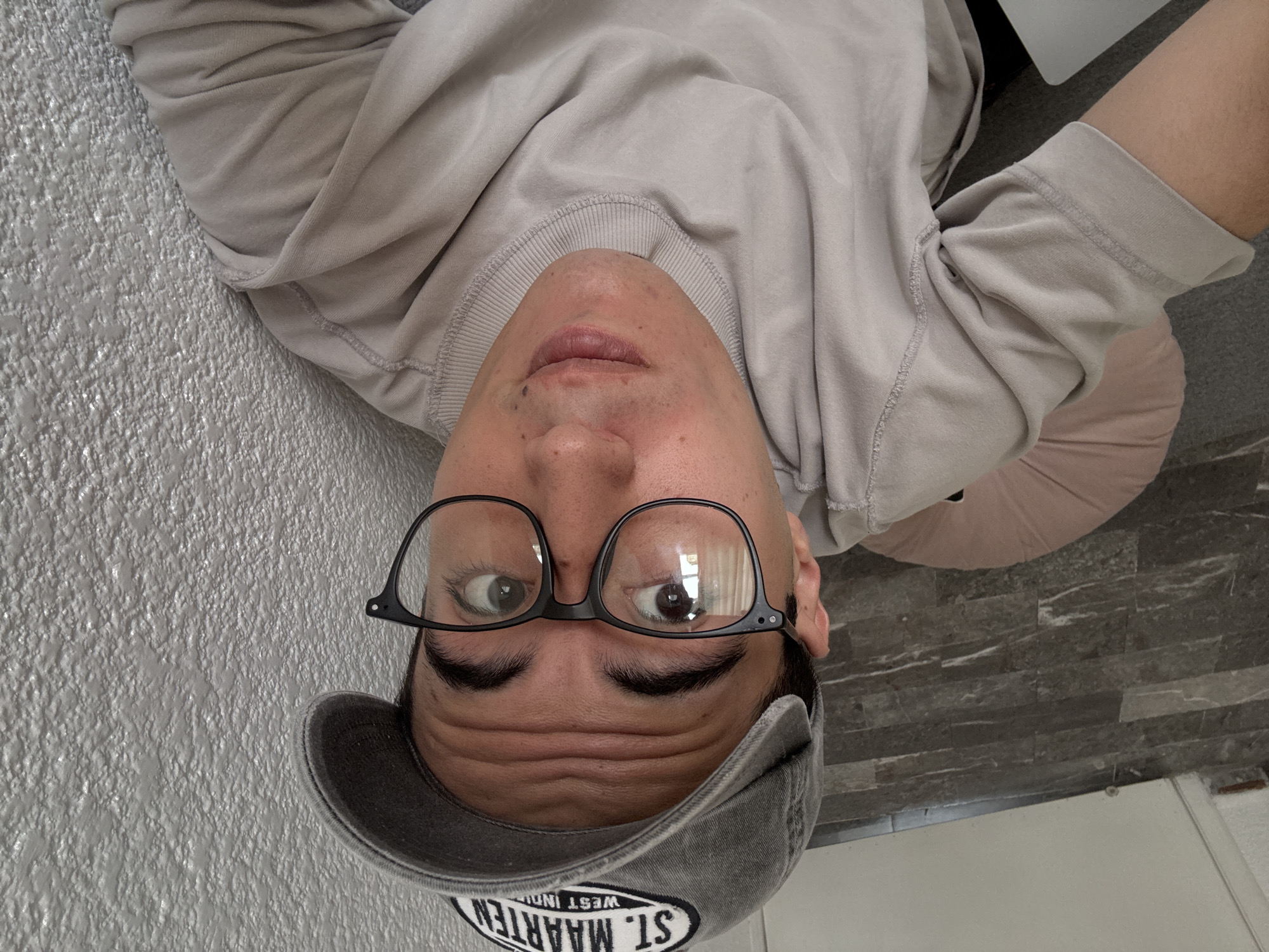 A close-up, high-angle selfie shows a person wearing a gray baseball cap and glasses, looking directly at the camera. The person has dark hair and appears to be wearing a light-colored t-shirt. Behind the person, the wall has a textured, bumpy surface, suggesting it is a stucco or concrete wall. The floor appears to be tiled with dark gray, rectangular tiles. A light pink, circular object, possibly a cushion or yoga ball, is visible to the left of the person. The scene does not contain any identifiable landmarks or clues that would place it in Puebla, Mexico.