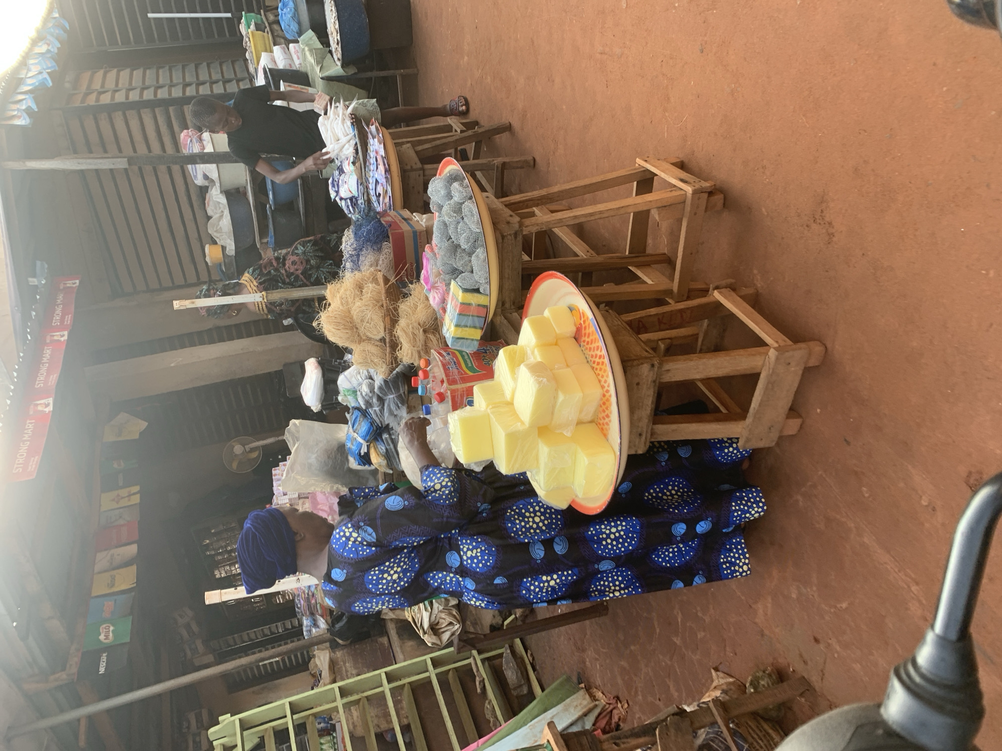 The image depicts an outdoor market stall in Shagamu, Nigeria. In the foreground, a woman wearing a blue and black patterned dress and a blue headscarf stands to the left, facing a display of yellow soap bars. To her right, a young man in a black t-shirt is seen attending to a pile of white items on a tray, possibly fish or dried goods.
The stall is covered by a corrugated metal roof supported by wooden beams. Various items are displayed on wooden tables and stands. To the left, a green ladder is leaning against a structure. Behind the stall, a fan is visible, and shelves are lined with different products, including bags and bottles. The ground is covered in reddish-brown earth, typical of the region. The scene suggests a bustling marketplace atmosphere.