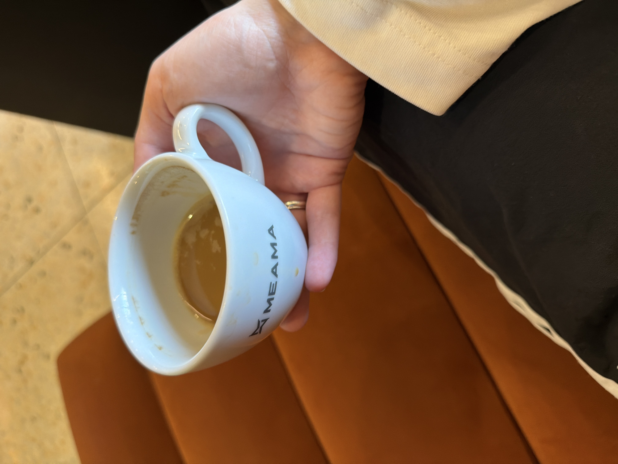 A person's hand holds a white coffee cup in a close-up, slightly angled shot. The cup contains a small amount of light brown liquid, consistent with the dregs of coffee or espresso. A gold-colored ring is visible on one of the person's fingers. The person is wearing a black garment with a distinctive white cuff or stripe, visible along the right side of the frame. The word "MEAMA" is printed vertically in black capital letters on the side of the cup, accompanied by a small, abstract logo.
The background features two distinct surfaces: a warm, orange-brown upholstered surface, possibly a chair or sofa, to the right and bottom, and a lighter, mottled surface, likely a table, in the upper left. The setting appears to be indoors, under ambient lighting. While the image itself does not offer specific geographical cues, the provided context indicates the location is Tbilisi, Georgia.