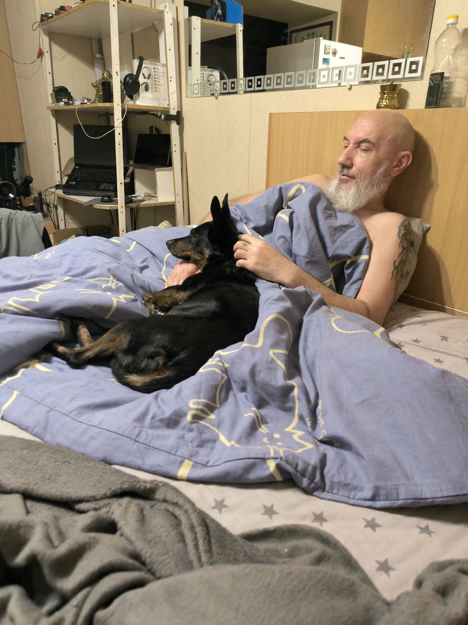 In an indoor setting, possibly a bedroom, a bald man with a grey beard is lying in bed. He is partially covered by a light blue duvet with a yellow pattern. A small black and brown dog is lying next to him, also under the duvet, with its head resting on the man's arm. The man appears to be looking to the side, with his eyes closed or nearly closed. The background includes a wooden headboard, shelving units with various items including electronics and what appear to be framed pictures, and a mirror reflecting items in the room. The overall atmosphere suggests a moment of rest or relaxation. The provided image does not contain any identifiable information or features that would allow for its specific location within the city of Mykolaiv, Ukraine, or any other specific geographical location.