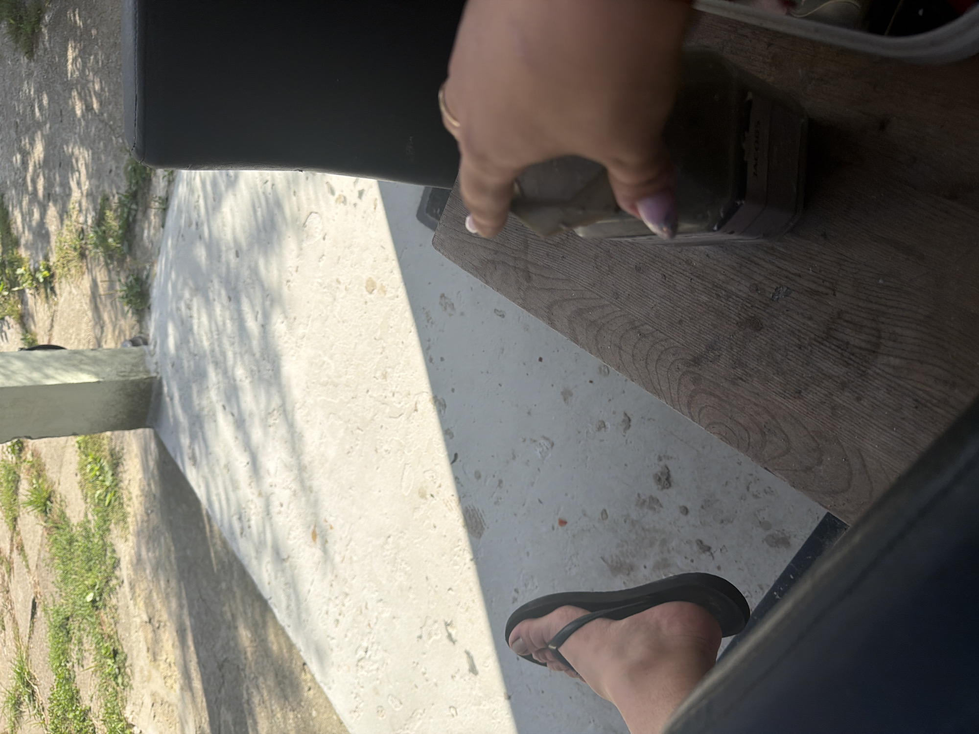 The image shows a close-up view of a person's hand and foot. The person is wearing a black flip-flop on their left foot, and their toes are visible. The hand is reaching towards a glass jar, which appears to contain some sort of green substance. A ring is visible on the person's ring finger. The scene appears to be outdoors, with concrete and grass visible in the background, suggesting a backyard or patio area. The lighting indicates it is daytime. The location is the city of Szczecin, Poland.