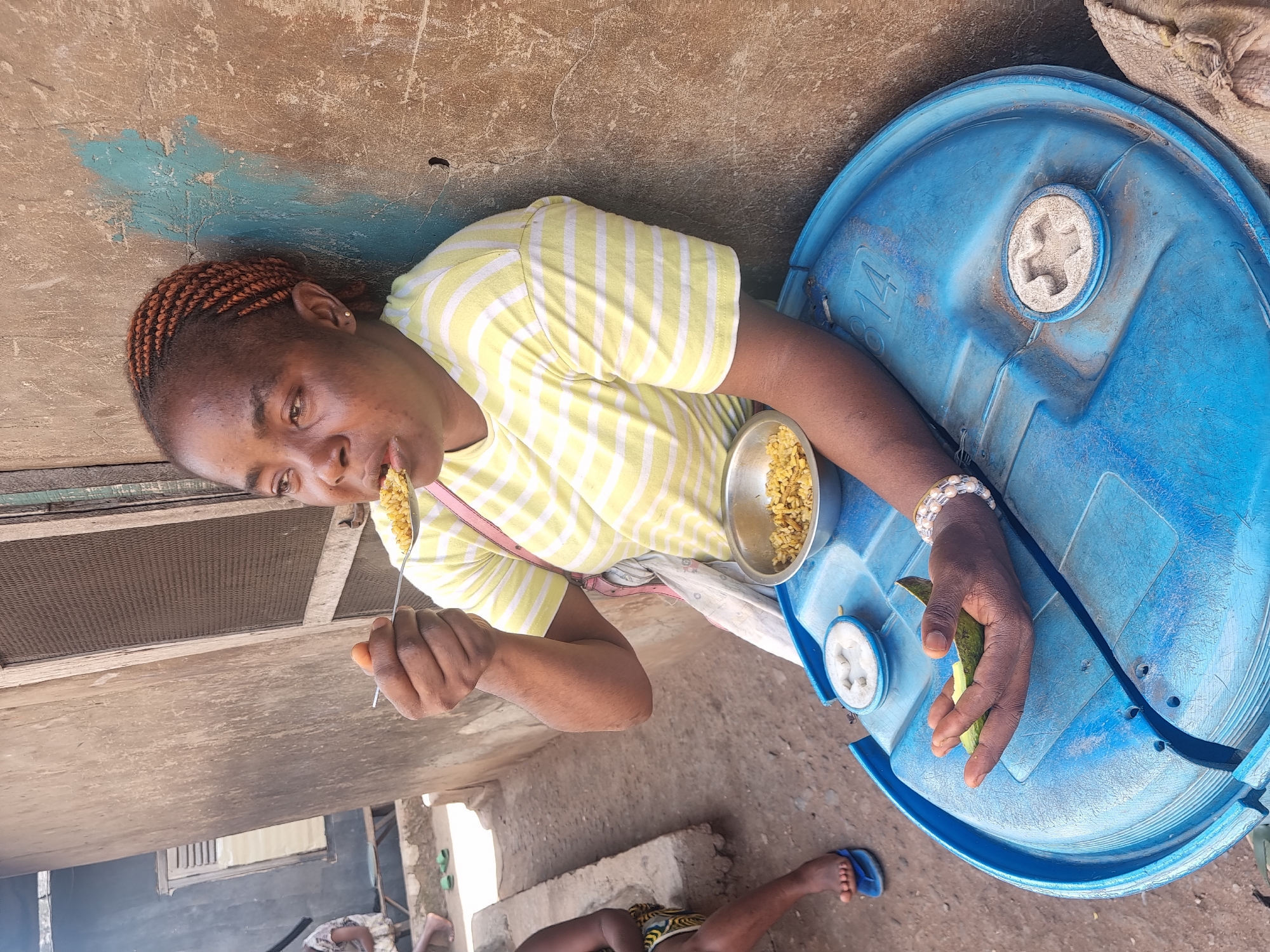 An outdoor, slightly rotated, overhead shot captures a woman eating, framed against a textured wall and ground. The main subject is a woman with dark skin and reddish-brown braided hair, wearing a yellow and white striped t-shirt. She is actively eating, holding a spoon with a spoonful of yellowish granular food, likely grains or rice, directed towards her mouth. A small metal bowl containing more of this food sits next to her.
Her left hand, adorned with a beaded white bracelet, rests on a blue plastic barrel. In this hand, she holds a piece of green, elongated fruit or vegetable. The blue barrel, a prominent object in the foreground, appears to be serving as a surface or makeshift table. Part of the number "814" is visible on its side.
The setting appears to be an informal outdoor or semi-outdoor space. The ground is a mix of concrete and dirt, showing signs of wear and cracks. The wall behind her is a rough, light brown concrete surface with patches of faded blue paint. In the background, hints of other simple structures, possibly a screened window or vent, are visible. At the bottom left, the leg and foot of another person, wearing a blue flip-flop, can be seen, suggesting others are nearby. The bright, clear lighting indicates it is daytime. The woman's expression is focused on her meal.