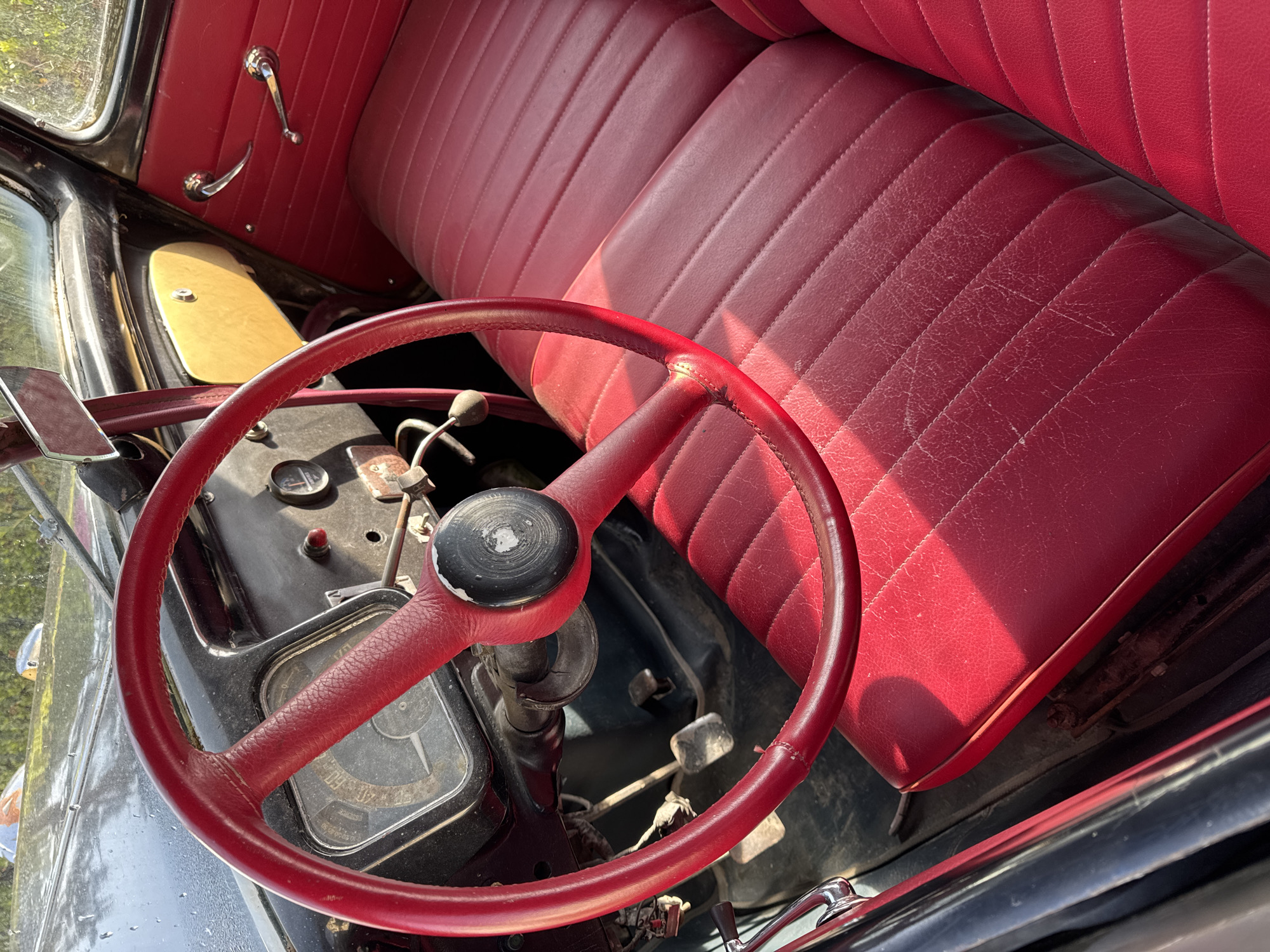 The image depicts the interior of an antique vehicle, likely a car or truck, in what is assumed to be Da Lat, Vietnam. The focal point is a large, red, circular steering wheel with a black center hub. Behind the steering wheel is a black dashboard with a gauge, a small red button, and various levers and switches. The seat is upholstered in red leather with vertical stitched channels. The image is taken from a low-angle perspective, looking up and across the interior of the vehicle. The environment is dimly lit. Parts of the exterior of the vehicle are visible, including a window and some of the bodywork. The overall impression is one of age and wear.