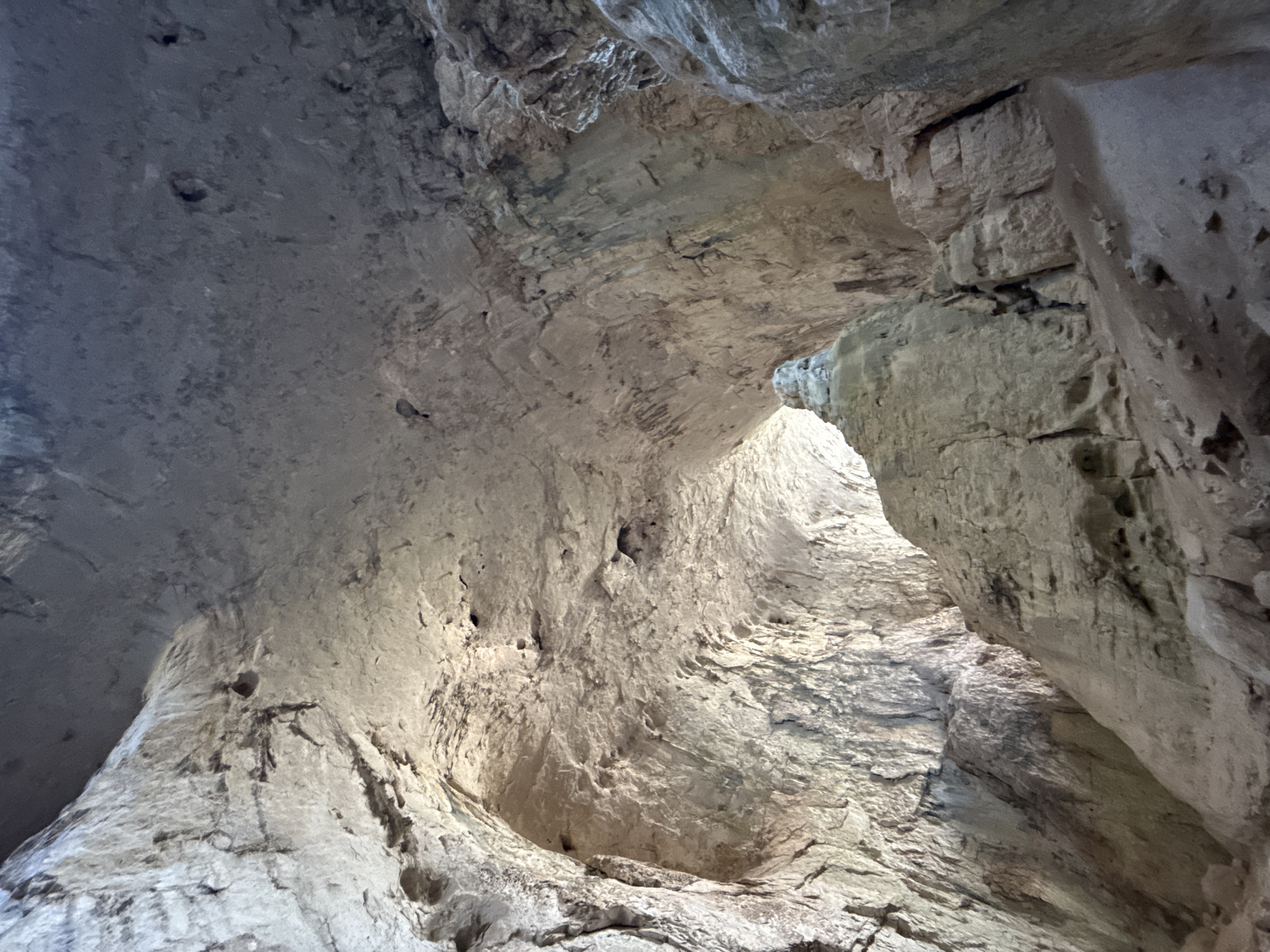 This is a photograph taken inside a cave in Karlukovo, Bulgaria, likely the Temnata Dupka cave. The image shows the interior of a large cavern with a rough, uneven ceiling and walls. The rock formations are primarily light brown and gray, with visible textures and striations. A large, weathered rock formation, possibly a stalagmite or a natural pillar, is prominent in the center foreground, casting a shadow. Natural light filters in from an opening in the distance, illuminating parts of the cave and creating contrasting shadows. The floor of the cave is covered in dirt and small rocks. The overall impression is one of a natural, ancient environment. There are no people or discernible man-made objects visible in the frame. The focus is on the geological features of the cave.