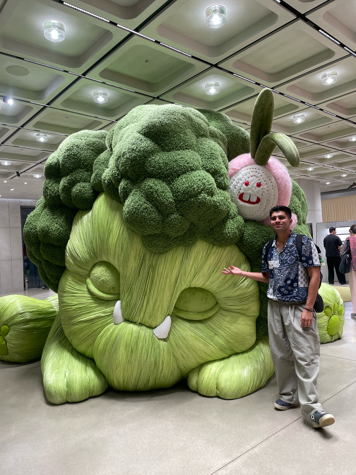 A man stands to the right of a very large, green, broccoli-like monster sculpture in a museum. The sculpture features a large, fuzzy green head resembling broccoli florets, with a smaller, round, white, bunny-like face with pink ears and red eyes nestled on top. The monster has large, bulbous green limbs. The man is wearing a patterned blue and white short-sleeved shirt, grey pants, and blue and white sneakers. He is smiling and gesturing towards the sculpture with his left hand. The background shows a modern ceiling with recessed lighting and a grid pattern. Other people are visible in the background, some walking and others standing. The environment appears to be an indoor exhibition space, likely a museum or gallery. The sculpture's placement in Seoul, South Korea, is not explicitly indicated in the visual information but is provided as context for the description.