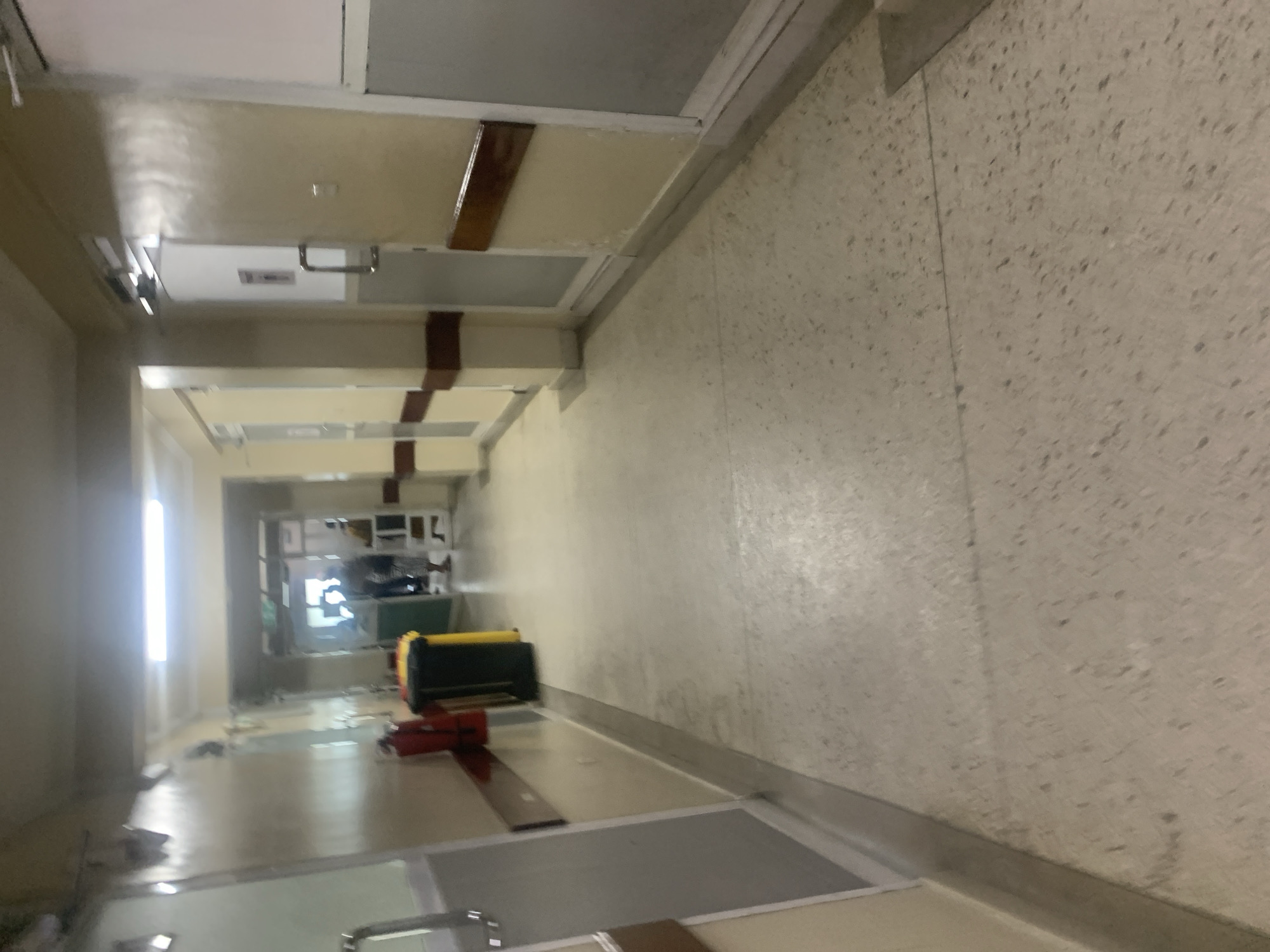 The image depicts an indoor hallway in what appears to be a building in Dar es Salaam, Tanzania, based on architectural styles and typical interior designs found in the region. The hallway has a beige, speckled wall on the right, and what looks like office-style ceiling panels overhead. Various objects are scattered down the hallway. Further down the hallway there are some lights. The precise nature of the objects and the building is not clear.
