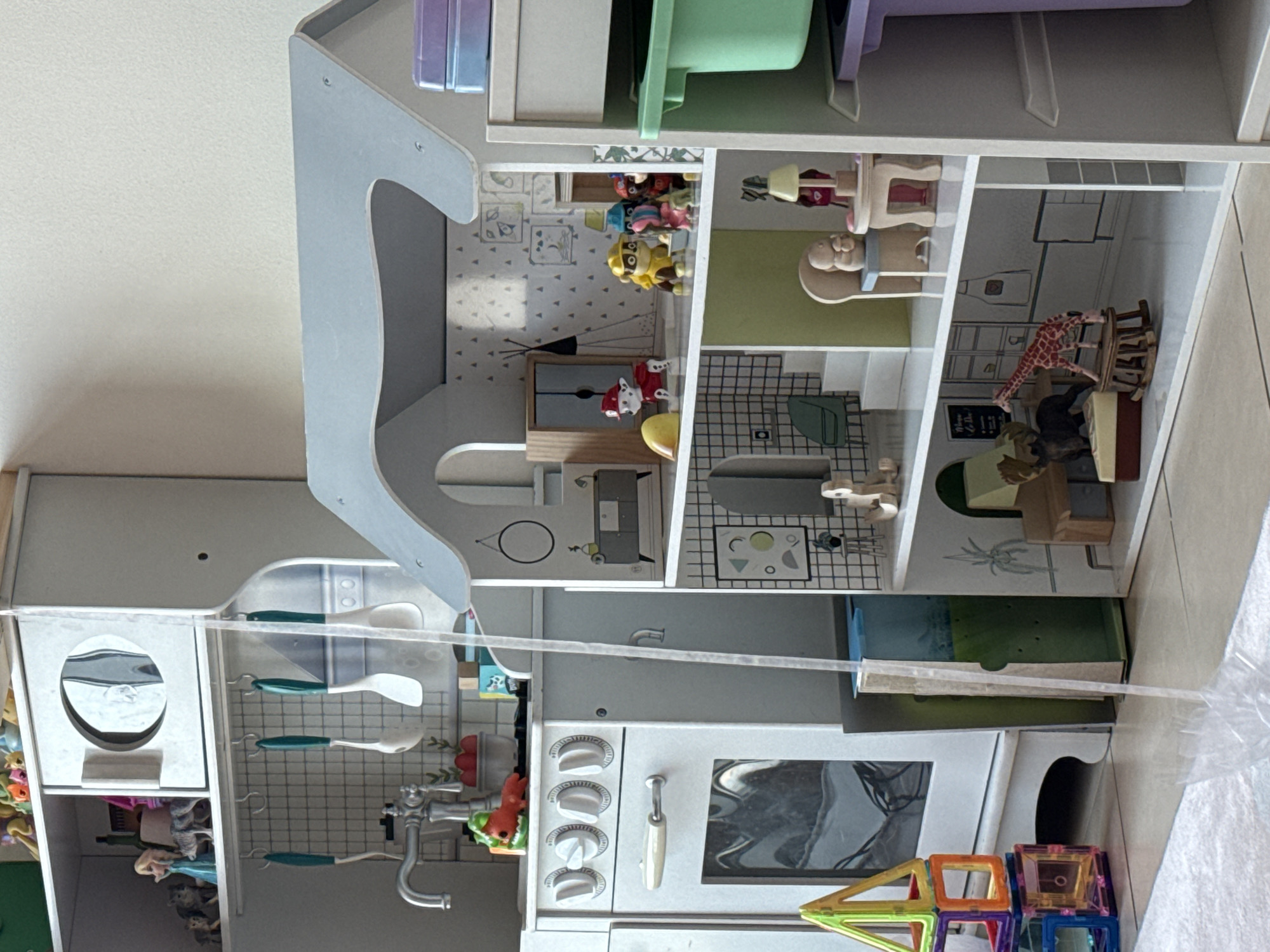 The image displays a collection of children's toys, including a dollhouse and a play kitchen, set against a plain wall and floor. The dollhouse, positioned to the right, features multiple levels with miniature furniture and figurines, including characters that appear to be from Paw Patrol and other animal figures. To the left of the dollhouse, a play kitchen set is visible, complete with a sink, faucet, oven, and various accessories like toy food and utensils. A stack of colorful magnetic building blocks is also present in the foreground. The overall scene suggests a child's playroom. There is no indication of the city of Dubai or the United Arab Emirates in the image.