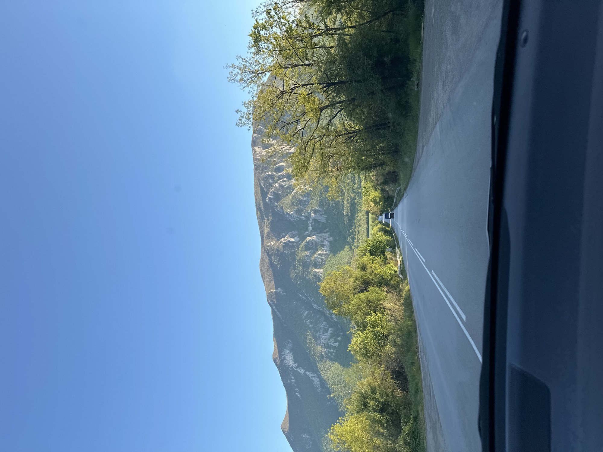 The image shows a view from a car driving on a road near Kravoder, Bulgaria. The road has two lanes separated by a double white line. A mountain rises to the left of the road, with green trees and vegetation covering its lower slopes. The sky above is clear and blue. Another car is visible in the distance on the road ahead. The car's interior is visible on the right side of the image.