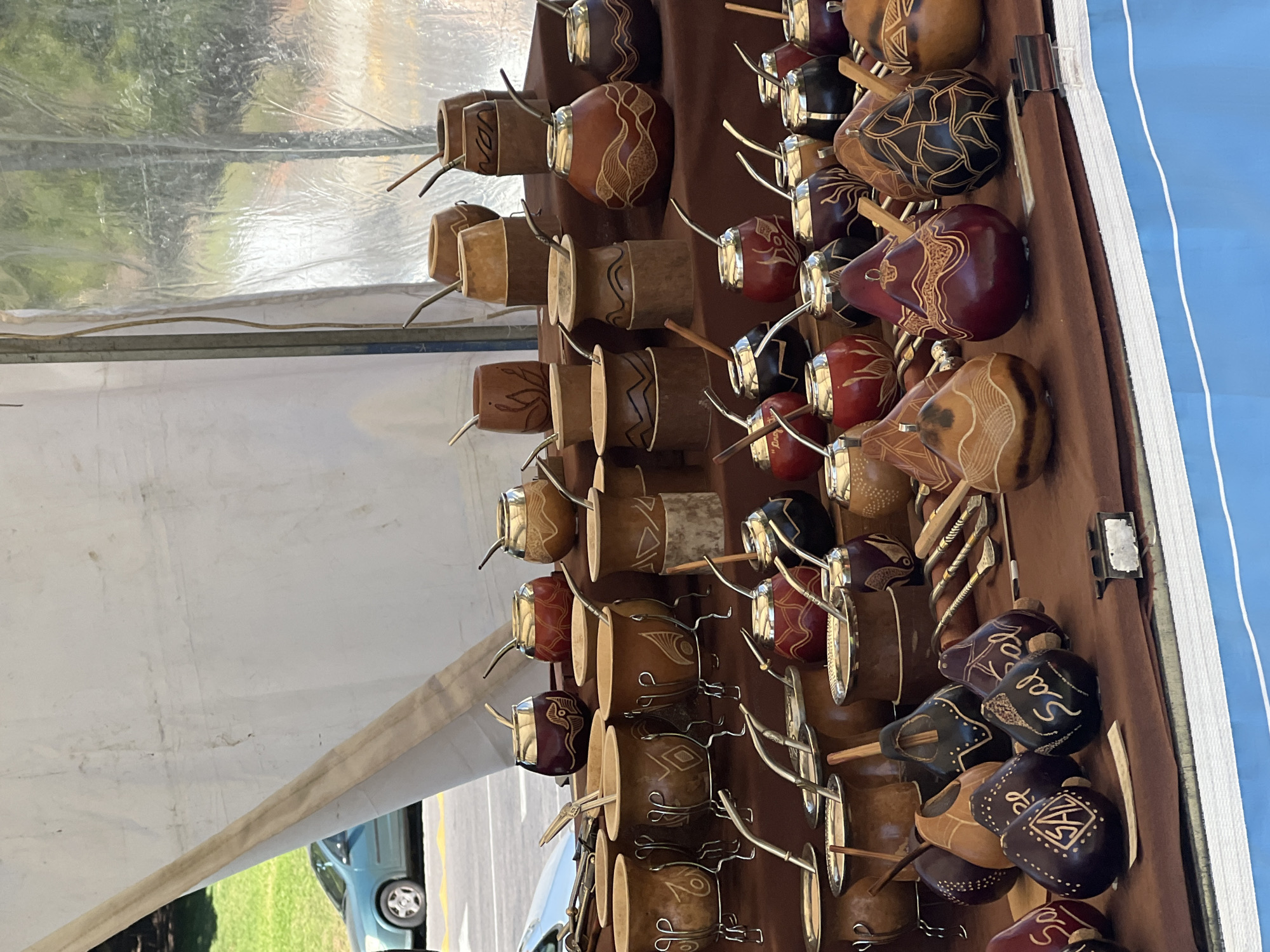 Rows of mate gourds are on display for sale, likely at an outdoor market in Buenos Aires, Argentina. The gourds vary in shape, size, color, and design. Each is fitted with a bombilla (metal straw). They are arranged on a brown, vertical surface, perhaps a stall or shelf. A car is parked outside and visible.
