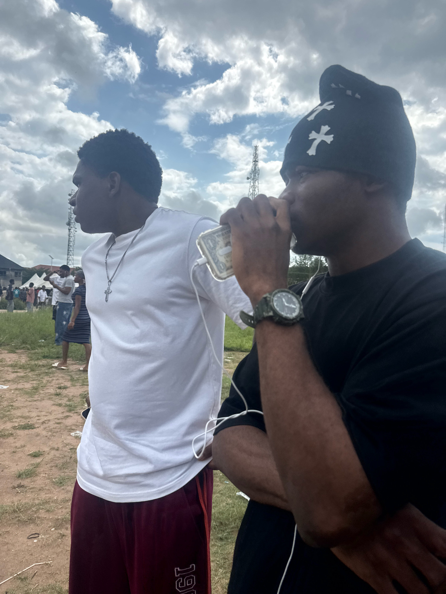In Ihiagwa, Nigeria, two young Black males stand outdoors under a cloudy sky. The male on the left wears a white t-shirt with a silver cross necklace and dark red sweatpants with white lettering. He is looking to his left. The male on the right wears a black beanie with white cross symbols and a black t-shirt. He is holding a phone to his mouth and looking to his right. He has a watch on his left wrist. In the background, several other people and tents are visible, along with tall communication towers. The ground is dirt and grass.