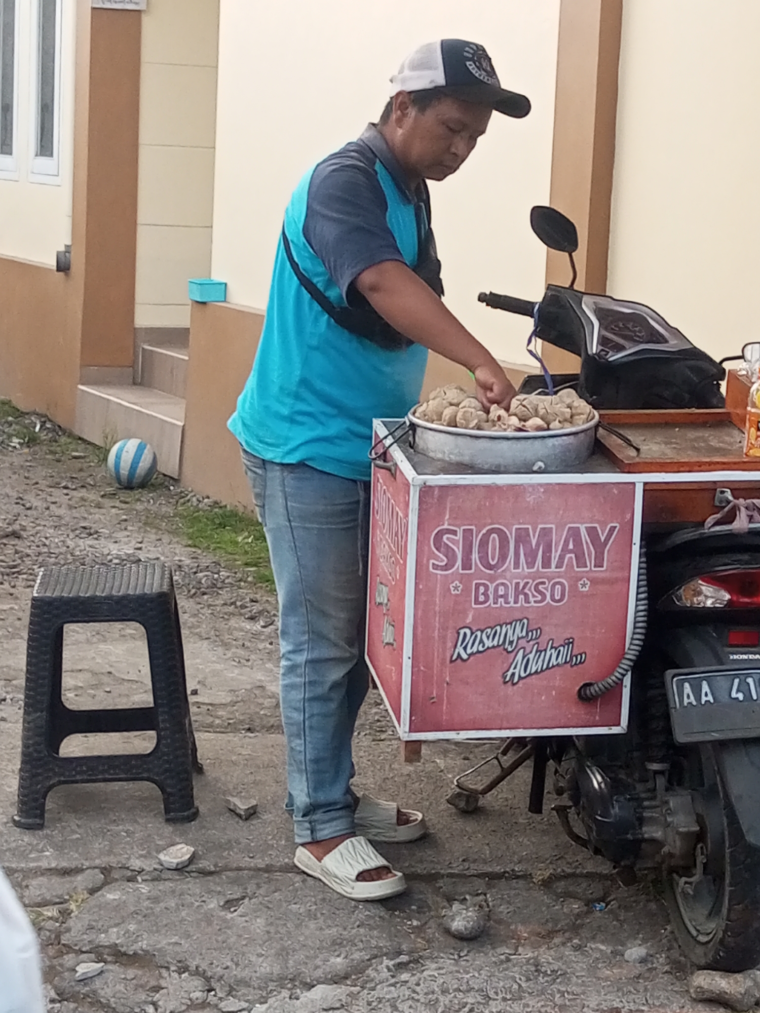 A man wearing a blue shirt, blue jeans, and a black and white baseball cap is standing next to a food cart attached to a motorcycle. He is in the process of serving food from a metal bowl filled with round, light-brown items. The food cart has a red side panel with white text that reads "SIOMAY BAKSO" and "Rasanya... Adahaii...". The motorcycle has a black seat and a black rear fender with a license plate that reads "AA 41...". A black plastic stool is positioned to the left of the man. In the background, there is a building with light-colored walls and brown trim. A blue and white striped ball is on the ground near the building's stairs. The ground is covered in a mixture of gravel and dirt. This scene appears to be outdoors, likely on a street or sidewalk in Pandansari, Indonesia.