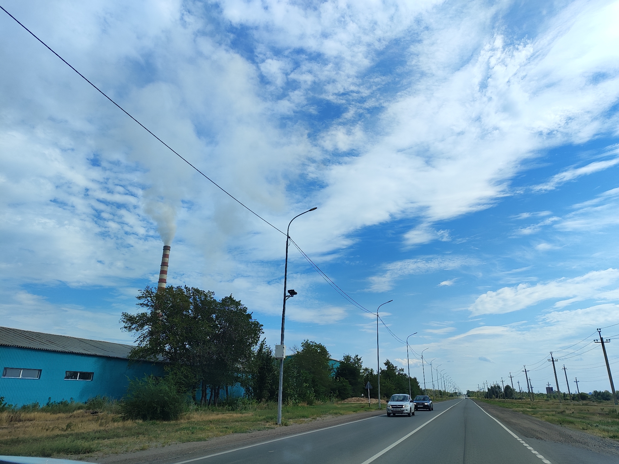 The image captures a view from a moving vehicle on a road, likely in Aksu, Kazakhstan, under a bright blue sky with scattered white clouds. To the left, a long, teal-colored industrial building with a grey roof is visible. Adjacent to the building, a tall, striped industrial chimney emits a plume of white smoke. A dense line of trees and bushes runs along the side of the road. Several streetlights and utility poles line the road, with power lines stretching across the frame. On the road, two cars are driving away from the camera: a silver car in the left lane and a darker car behind it in the right lane. The road itself has white lane markings. The surrounding landscape appears dry and grassy.